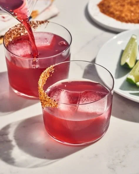 Two glasses of pink cocktails with rim sugar, ice, and lime wedges on a white surface with a plate of tacos and guacamole in the background.