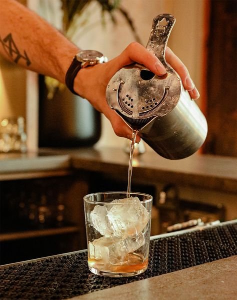 A person is pouring a drink with ice from a metal cocktail shaker into a glass on a bar counter.