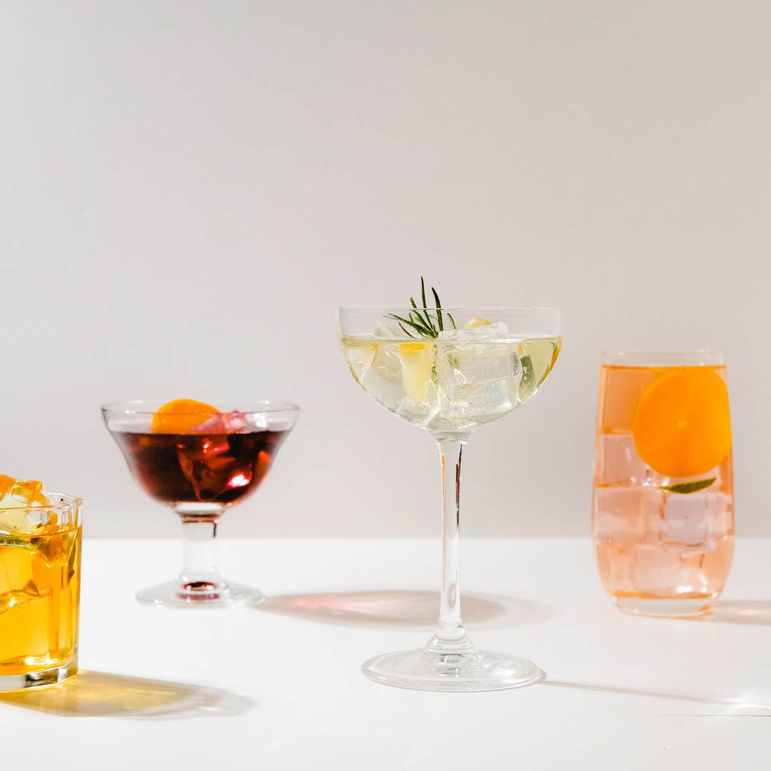 Four colorful cocktails on a white surface against a plain background, including a yellow drink in a short glass, a red drink in a coupe glass, a clear drink with ice and a sprig of rosemary in a coupe glass, and an orange drink with a slice of orange in a tall glass.