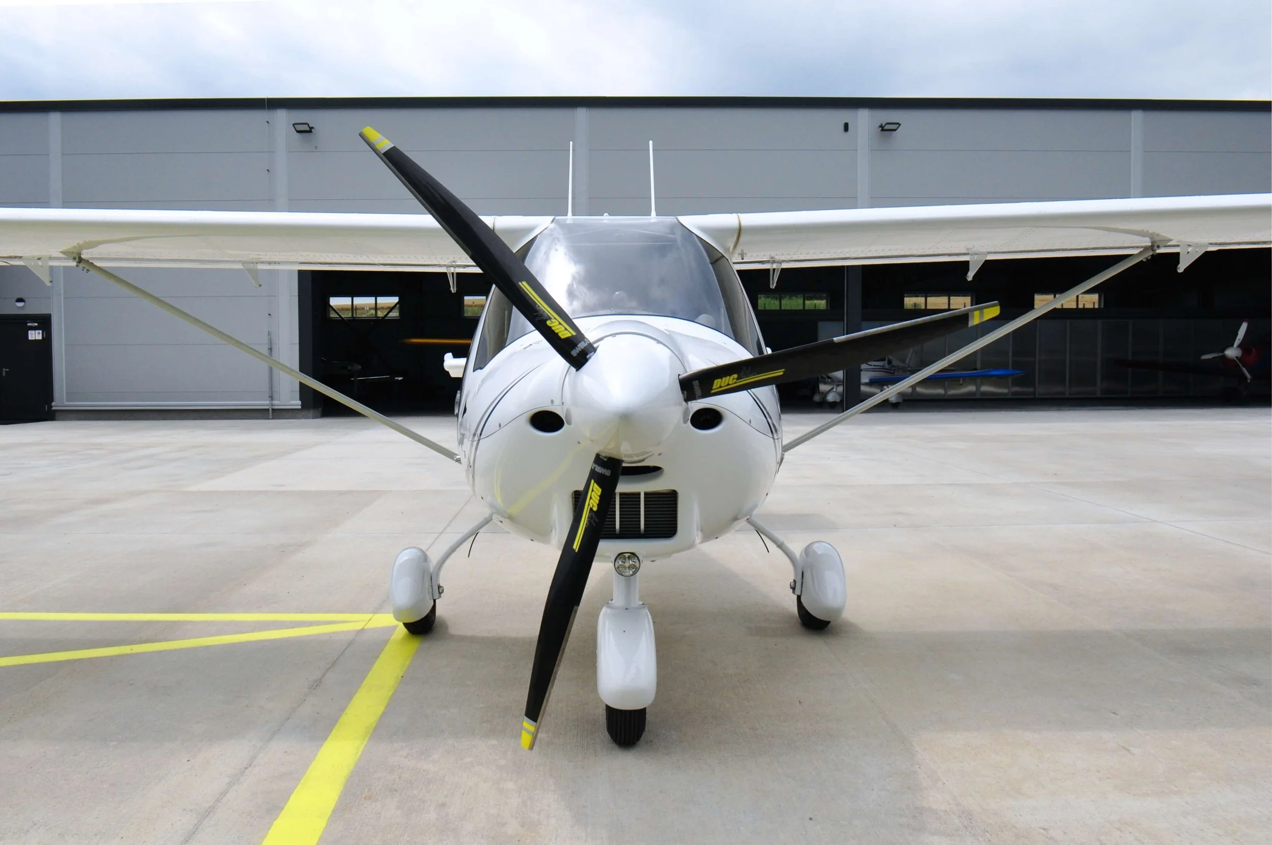 Kleines weißes Flugzeug mit dunklen Propellern vor einer Hangar-Gebäude auf Betonstrasse.