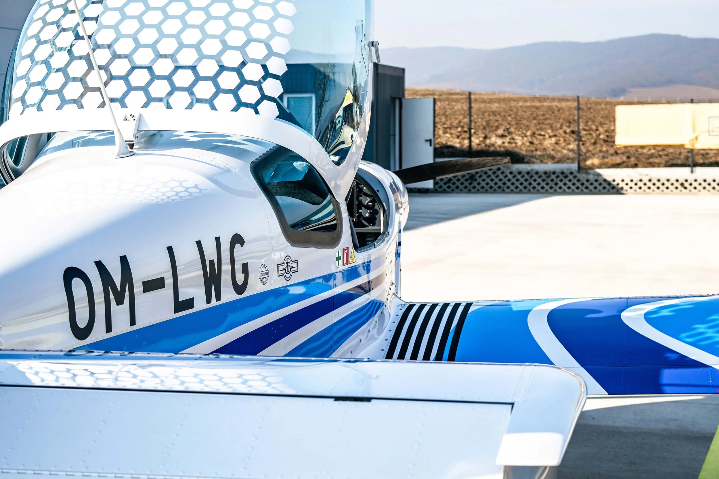 Ein Rennflugzeug mit blauer und weißer Bemalung, das auf einem Pipapo steht, mit Sicht auf Cockpit und Flügel, im Hintergrund eine Wüsten- und Berglandschaft.