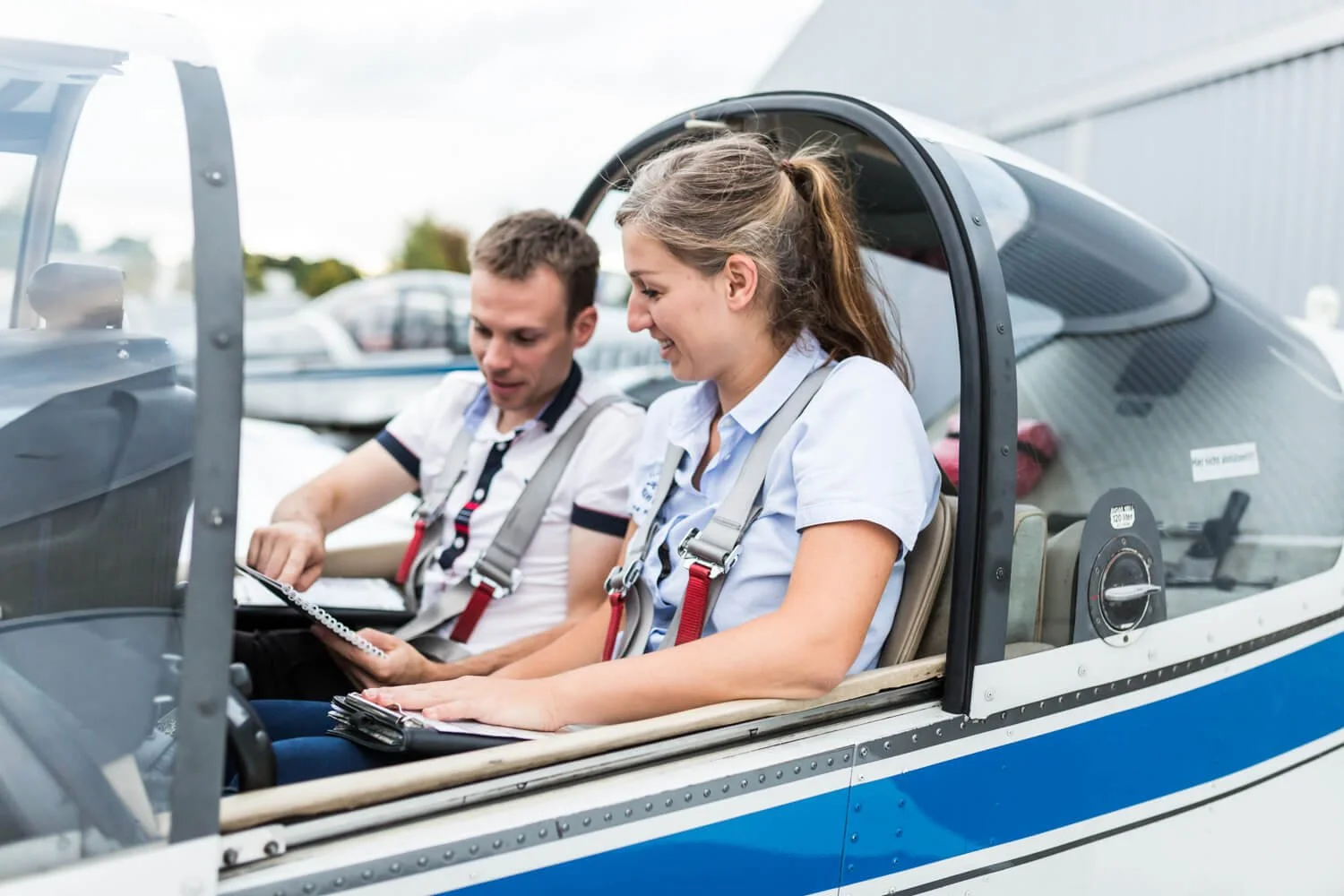Ein Mann und eine Frau sitzen in einem kleinen Flugzeug, beide tragen Sicherheitsgurte und scheinen sich auf einen Flug vorzubereiten, während sie lachen und sich unterhalten.