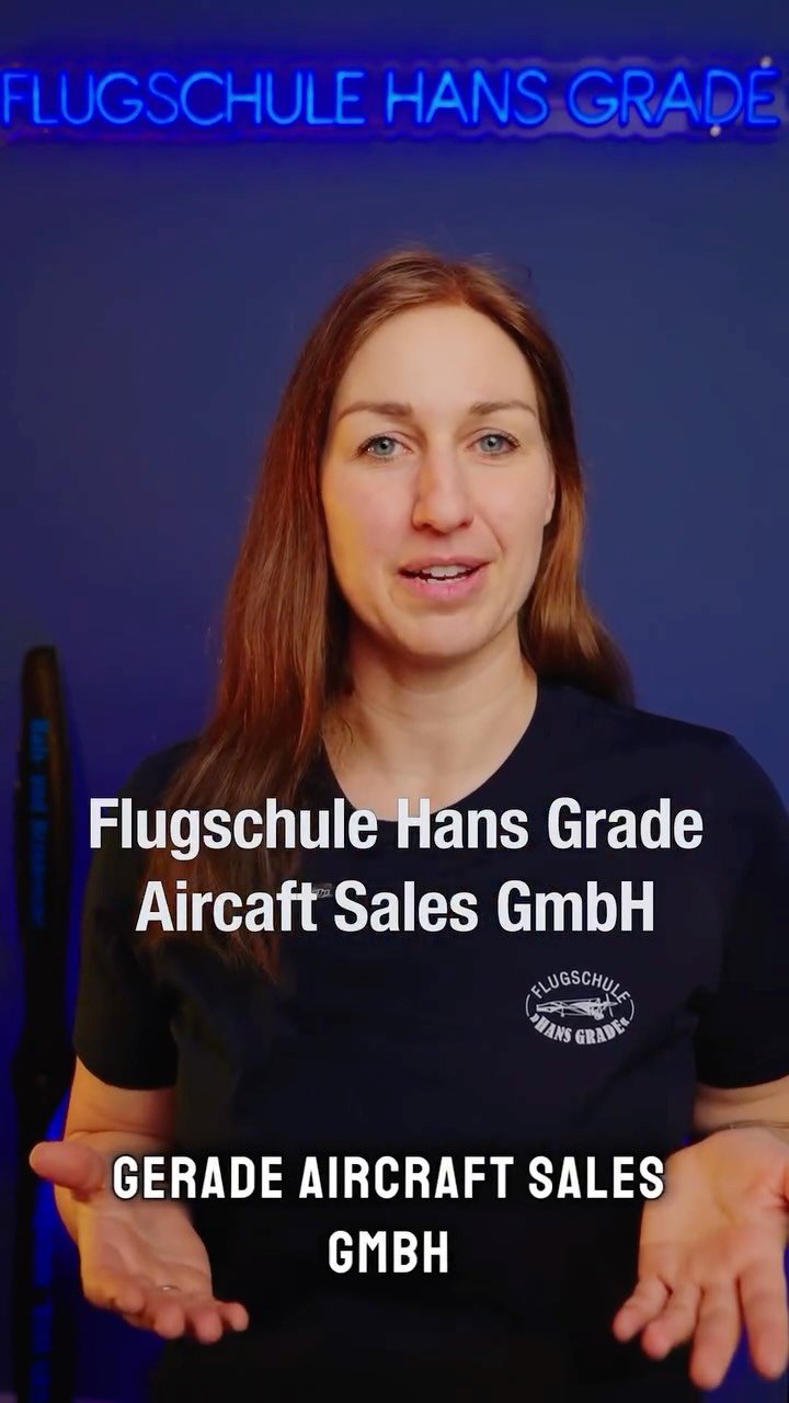 Frau steht vor einem blauen Hintergrund mit dem Neon-Schriftzug 'Flugschule Hans Grade'. Sie trägt ein schwarzes T-Shirt mit dem Logo der Flugschule und erklärt etwas, mit offenen Händen gestikulierend.