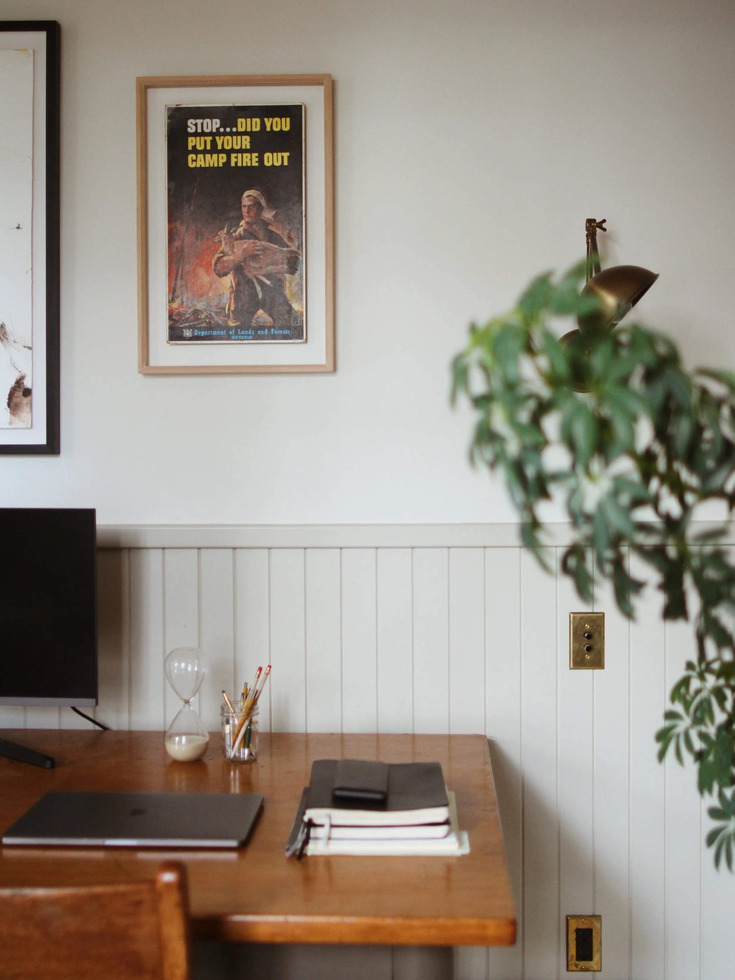 A home office desk with a closed laptop, notebooks, a phone, a glass jar with pens, and a small hourglass on a wooden table. The background features a white wall with a framed vintage poster that reads 'Stop... Did You Put Your Camp Fire Out' and a wall-mounted lamp.