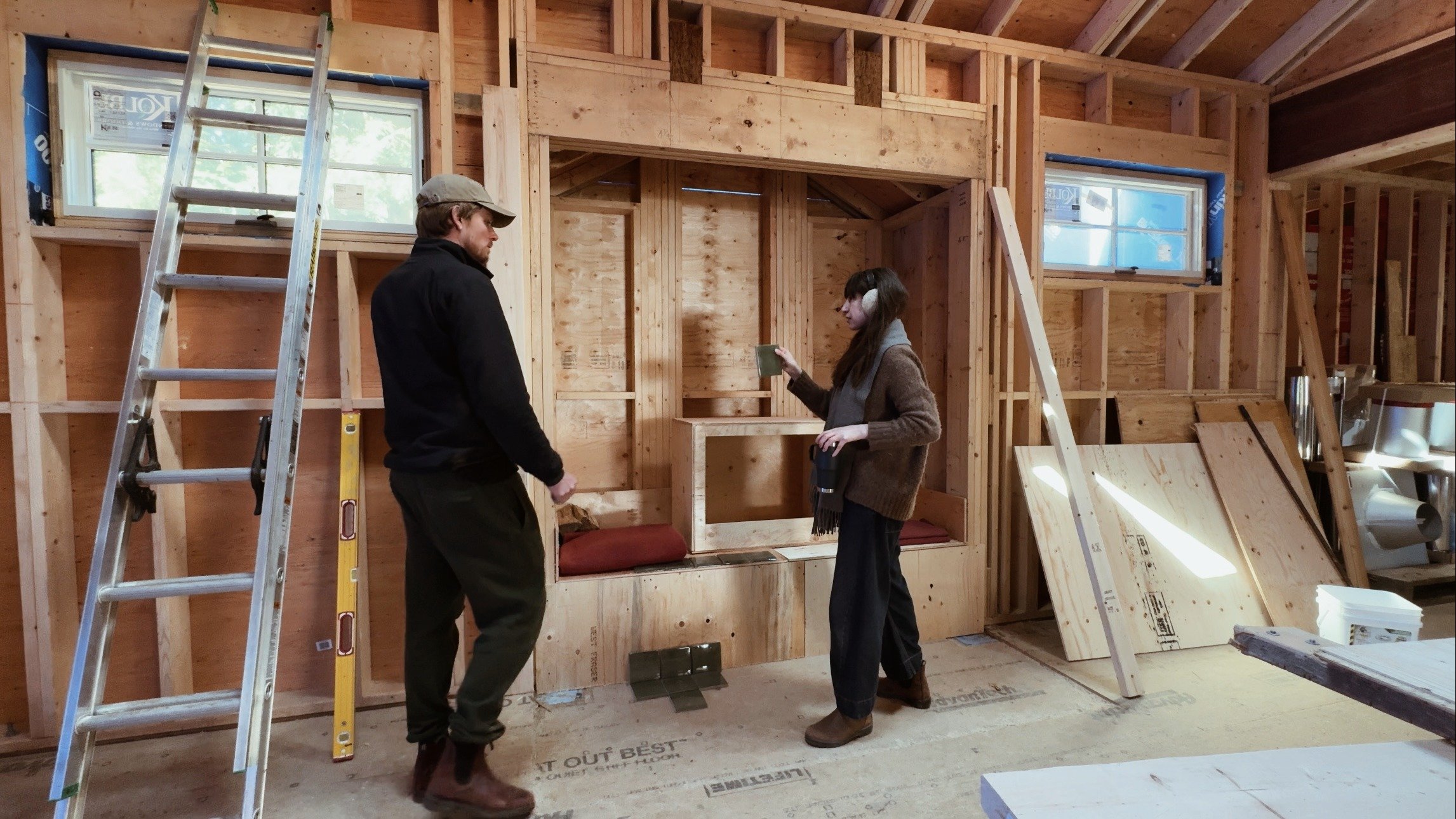 David and Lee stand in a large room with exposed framing, discussing the tile design for the central fireplace. There are building materials, levels and ladders in the space.