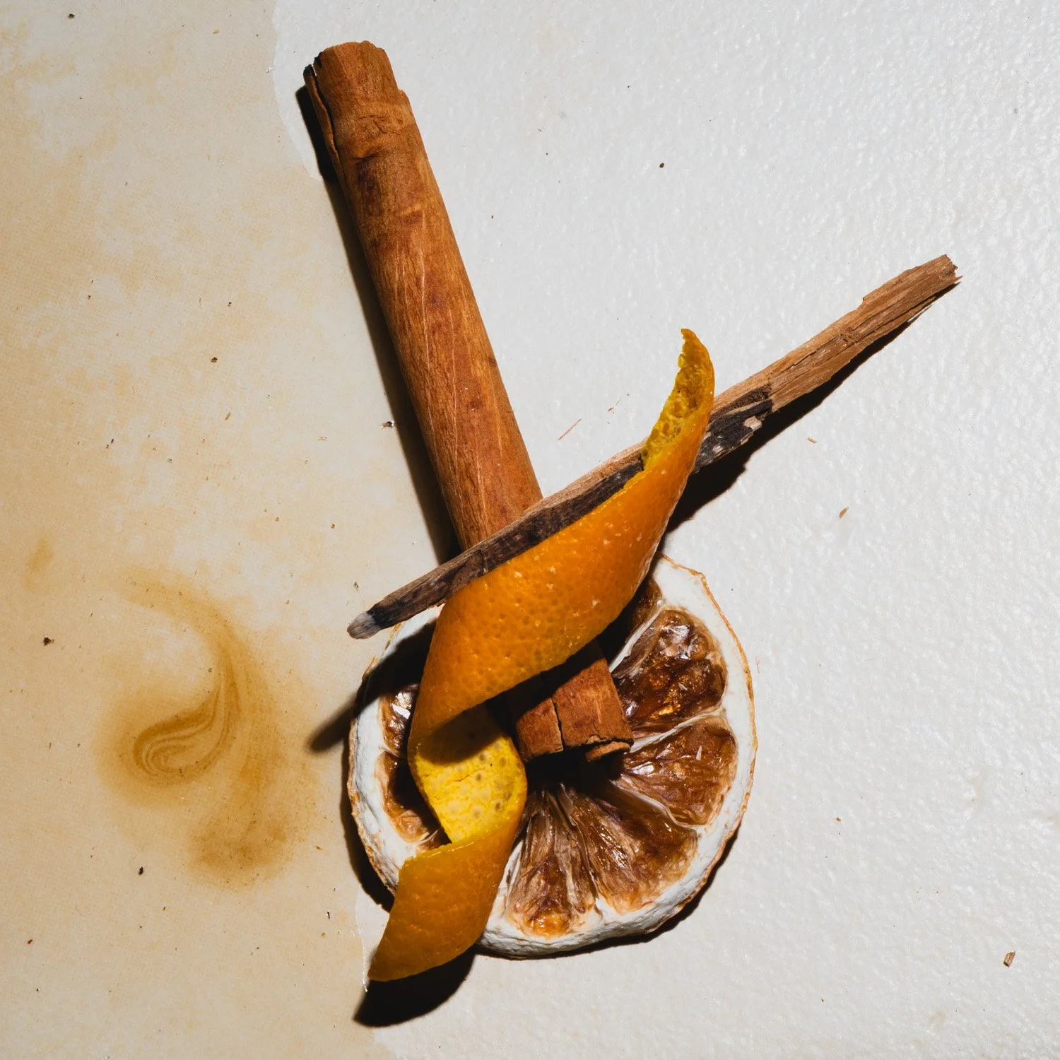 A dried lemon slice with orange peel, a cinnamon stick, a piece of dried brown fruit, and a cinnamon bark strip on a white surface.