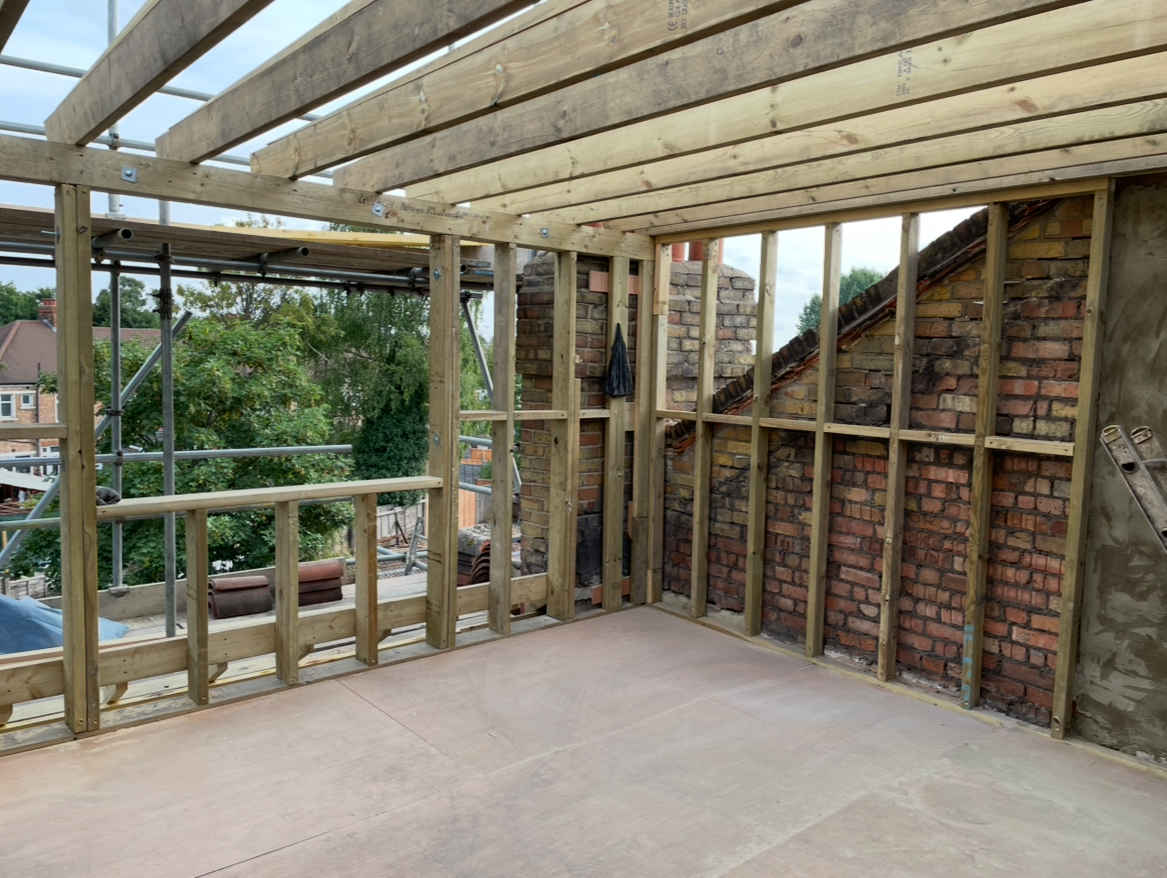Interior of a building under construction with exposed wooden framing and brick chimney, view of an outdoor neighborhood with trees and houses.