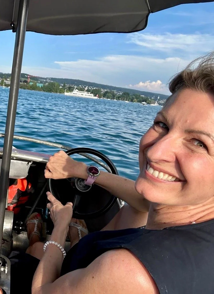 Eine Frau fährt in einem Boot auf einem See, lächelt in die Kamera, während die Wasseroberfläche und andere Boote im Hintergrund sichtbar sind.