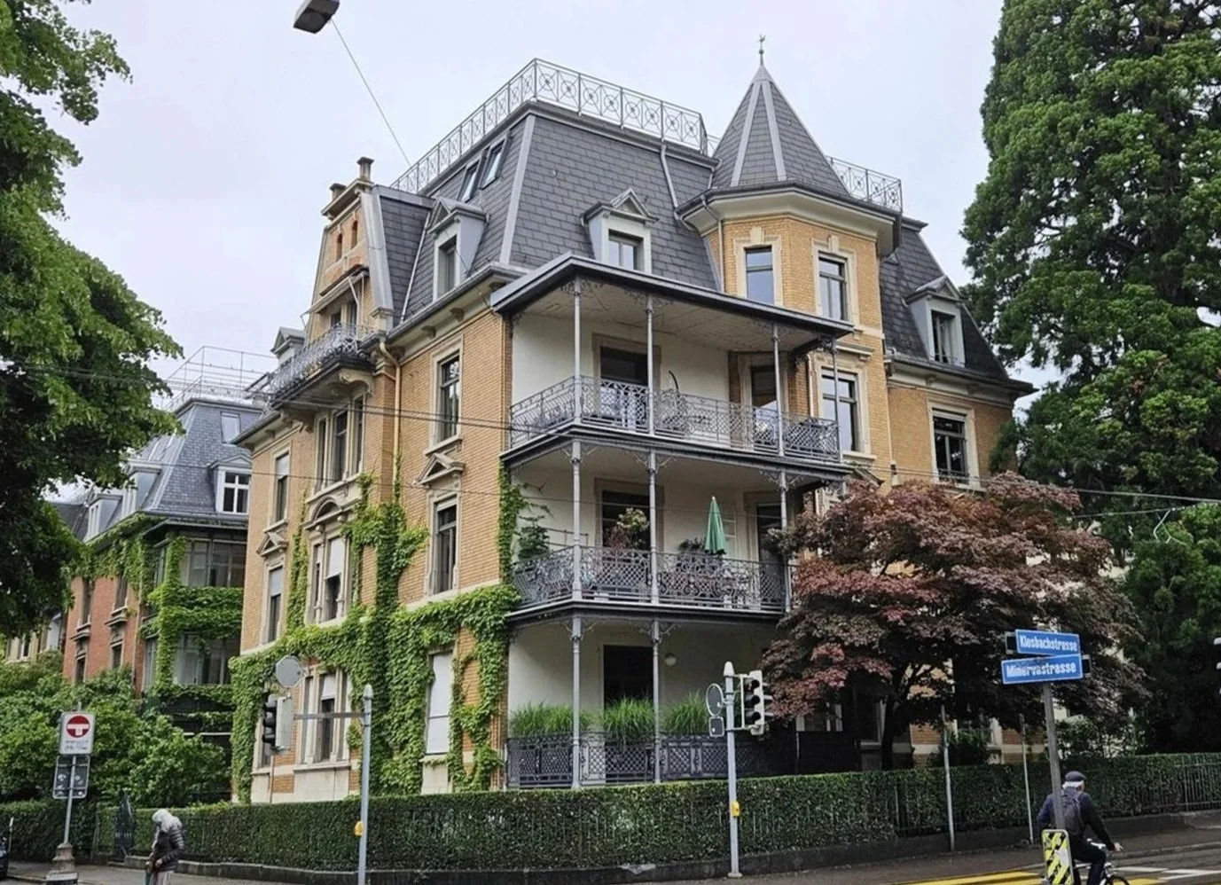 Ein mehrstöckiges, historisches Gebäude mit gelben Ziegelwänden, mehreren Balkonen mit Metallgeländern und einem spitzen Dach, umgeben von Bäumen, in einer Stadt.