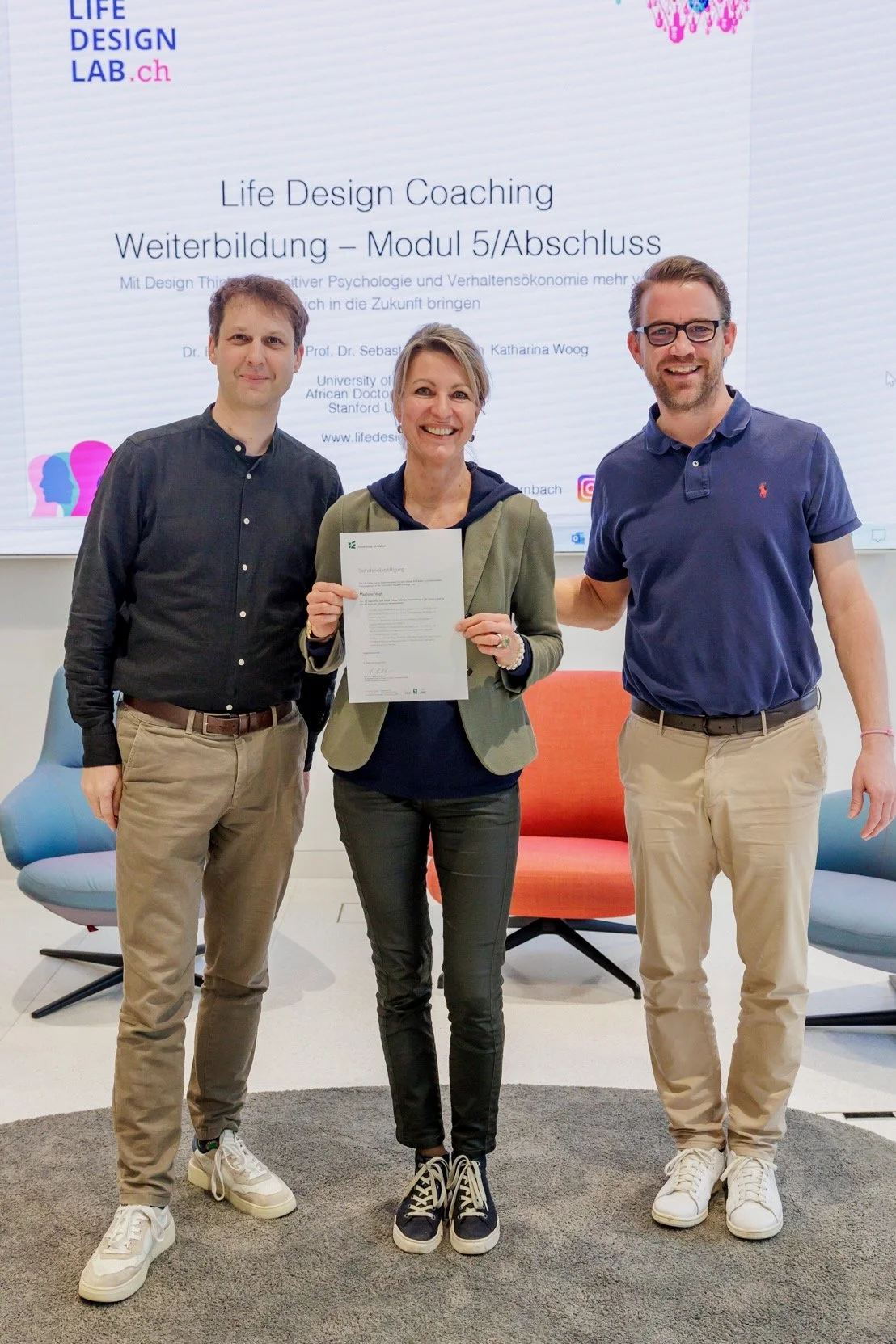 Drei Personen stehen vor einer Leinwand, die eine Präsentation mit Text und Logos zeigt. Die Frau in der Mitte hält ein Zertifikat. Es scheint sich um eine Abschlussfeier für eine Weiterbildung im Bereich Life Design Coaching zu handeln.