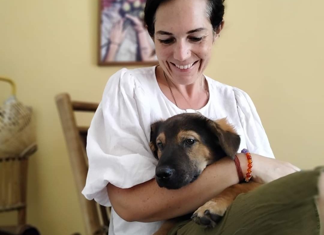 Woman smiling and holding a brown and black puppy indoors with a yellow wall, a framed picture, and wooden furniture in the background.