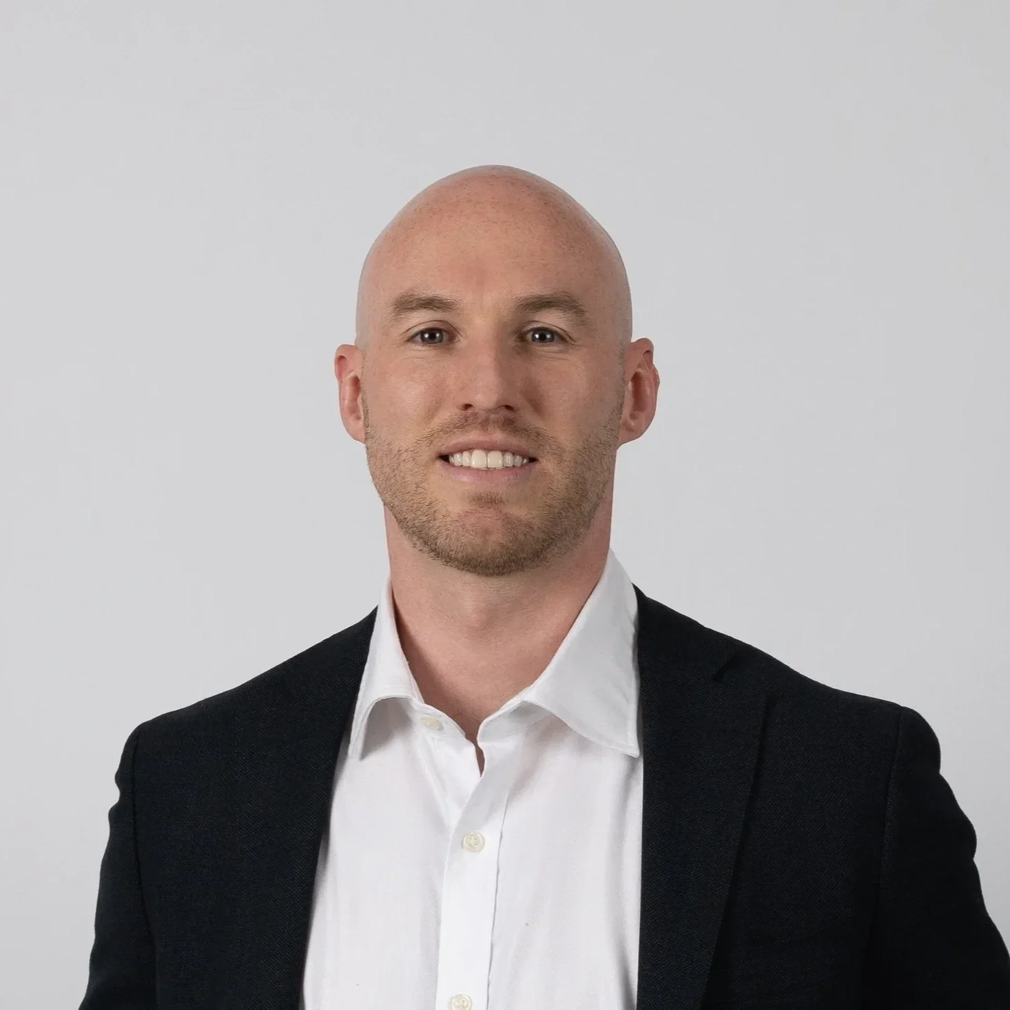 A man dressed in a white shirt and black blazer, smiling against a plain white background.