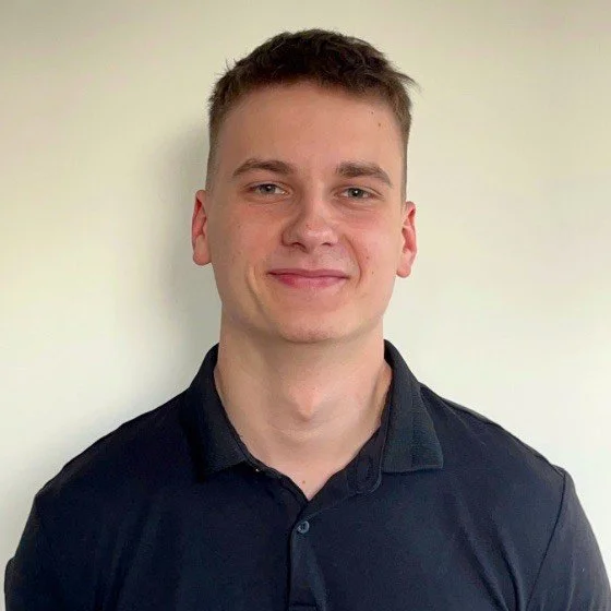 Portrait of a young man with short brown hair, wearing a black collared shirt, standing in front of a plain light-colored wall.