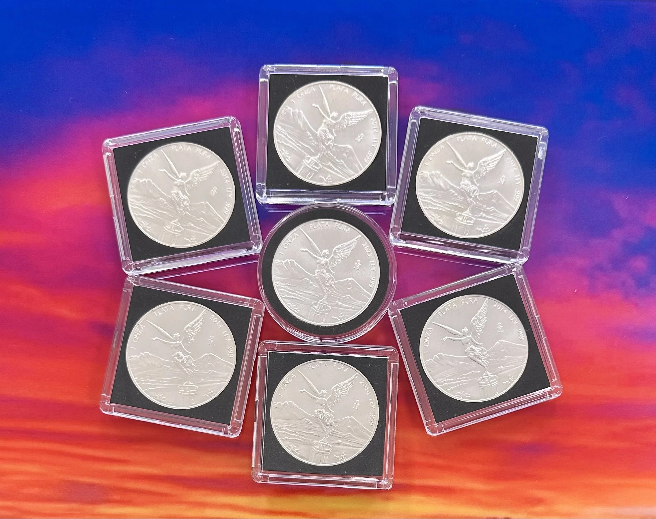 Seven silver coins in clear plastic cases arranged in a star shape on a colorful, sunset-like background.