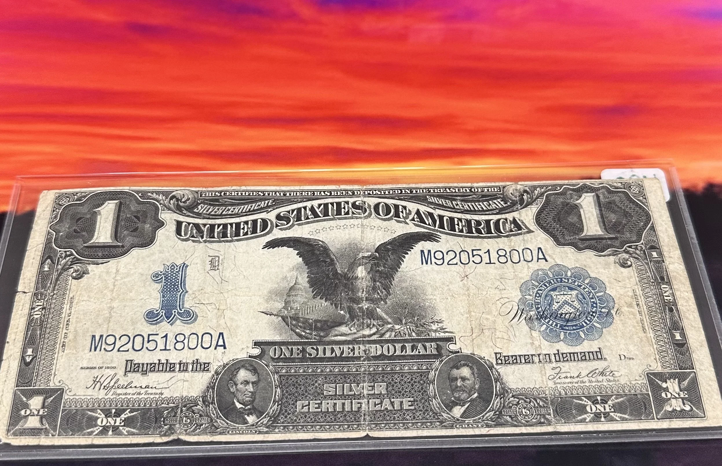 One dollar bill with eagle graphic in front of a vibrant orange and pink sunset sky.