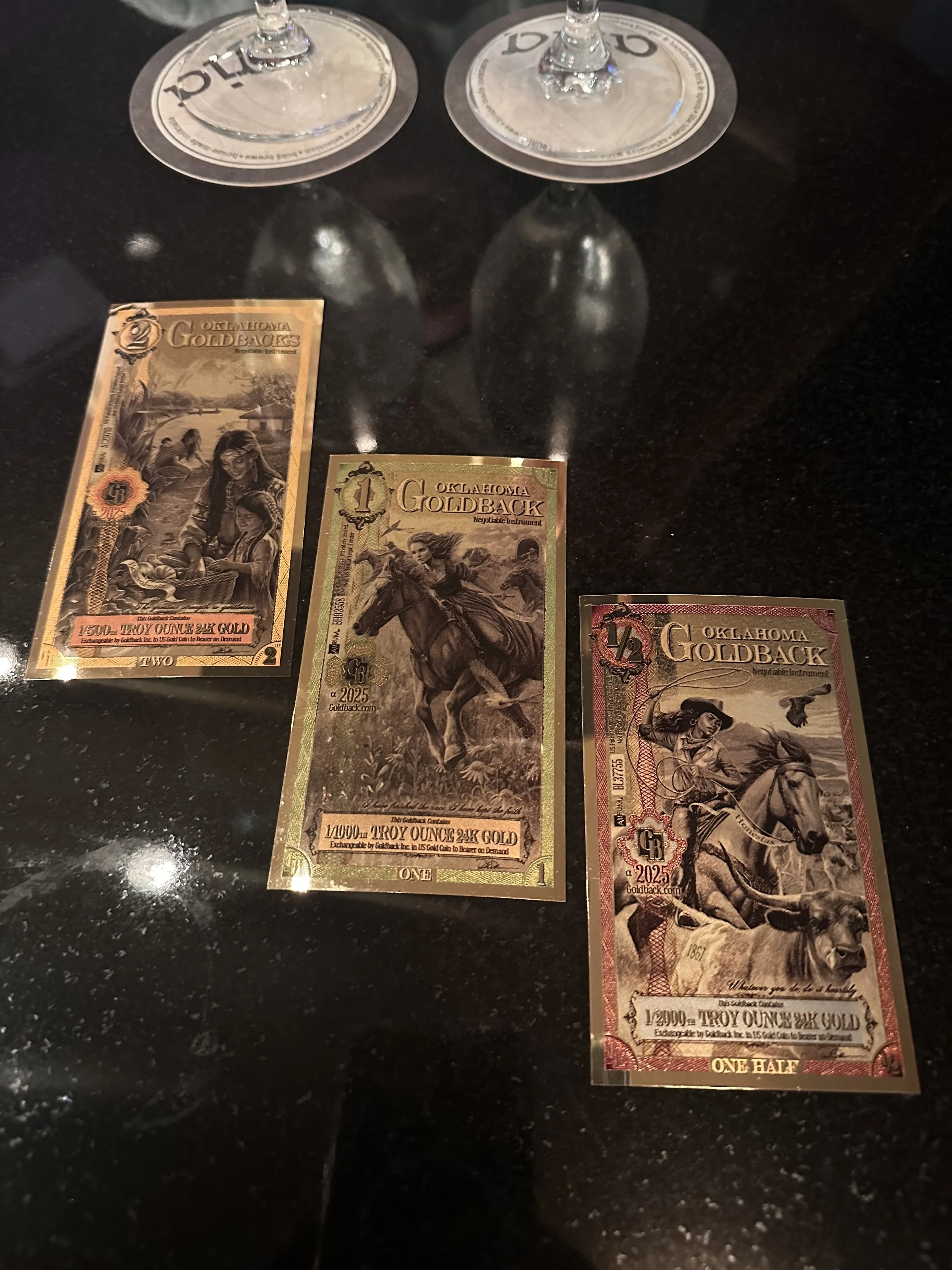 Three Oklahoma Goldback bills on a black surface, with two glasses upside down in the background.