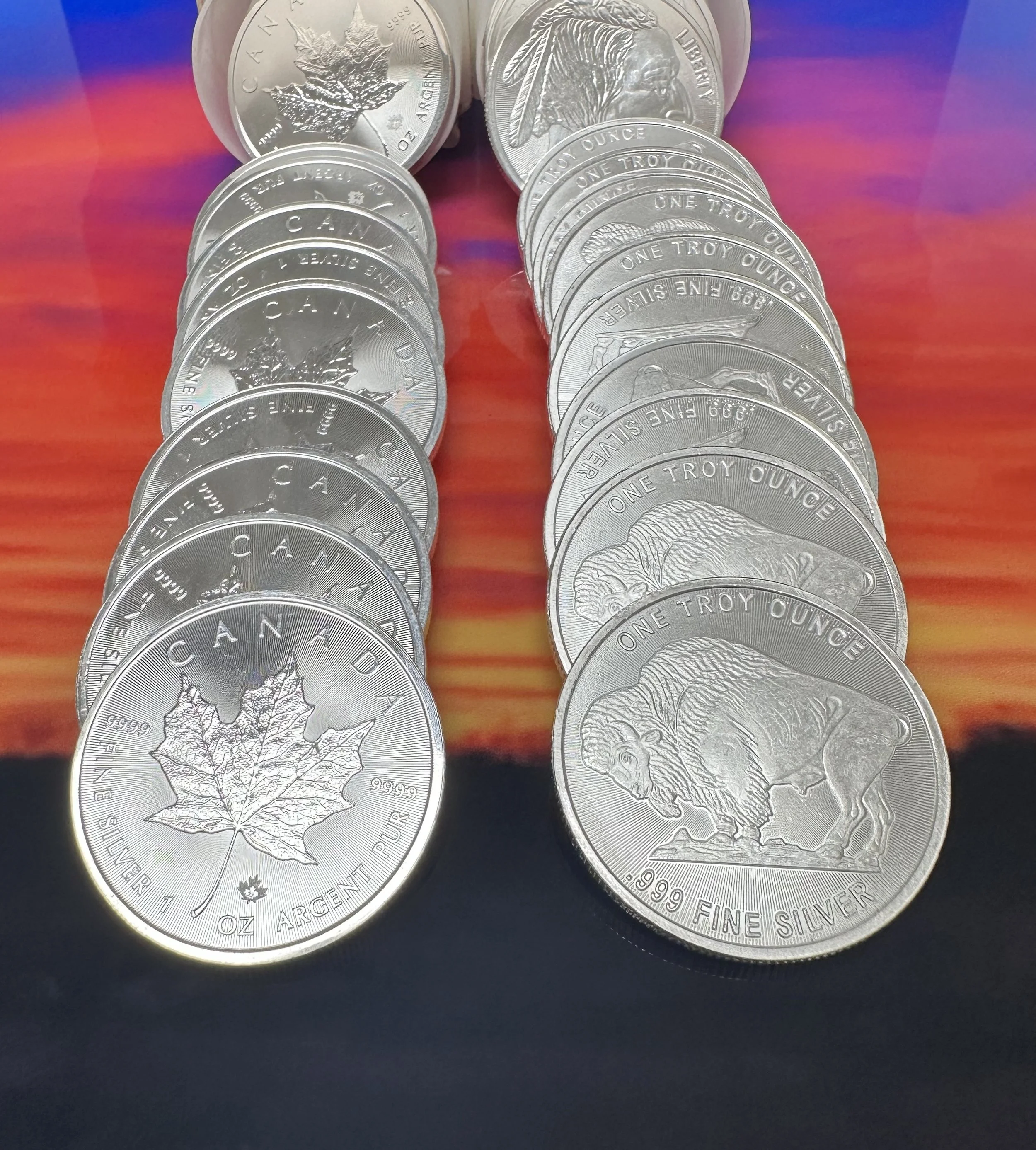 Arrangement of Canadian one-troy-ounce silver coins, featuring a maple leaf on some and a bison on others, on a colorful, sunset-like background.