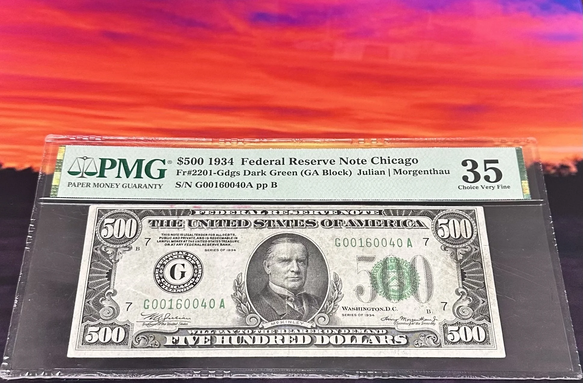 A 1934 Federal Reserve Note Chicago $500 bill in a protective plastic cover, labeled as 35 Choice Very Fine, against a vivid sunset background.