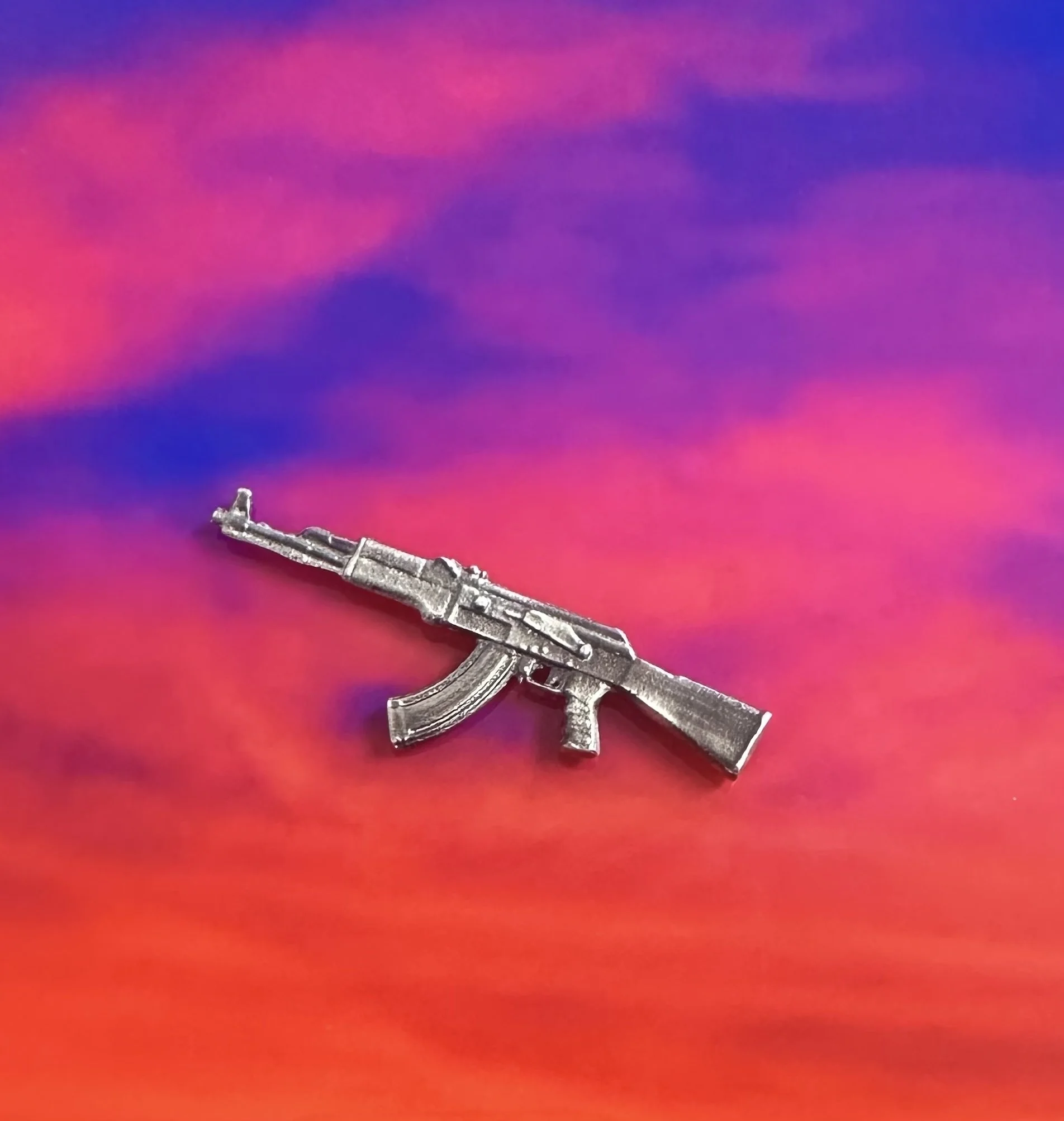 A small metallic silver gun-shaped pin on a colorful, swirling background of pink, purple, and orange.