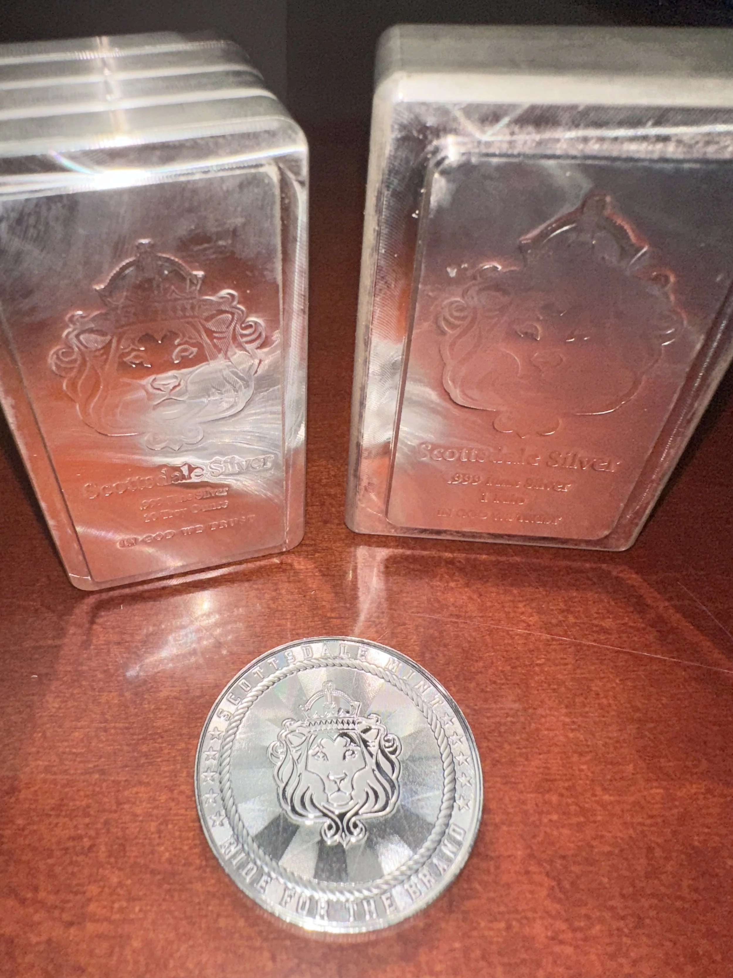 Two silver bars with engraved branding and a silver coin with an embossed lion and engraved text on a wooden surface.