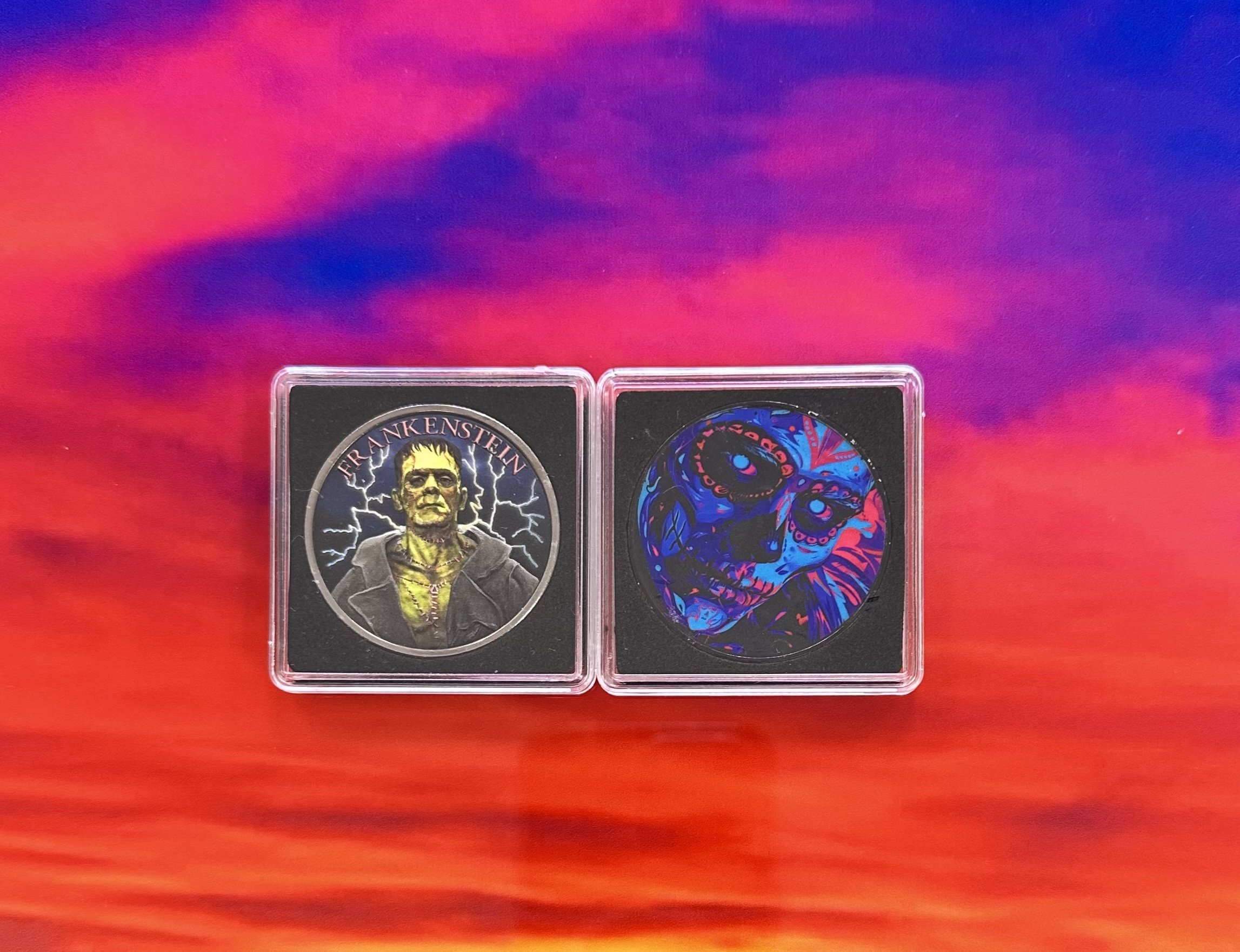 Two collectible cards in clear cases on a vibrant, colorful background. The left card features Frankenstein with lightning in the background, and the right card has a colorful skull design.