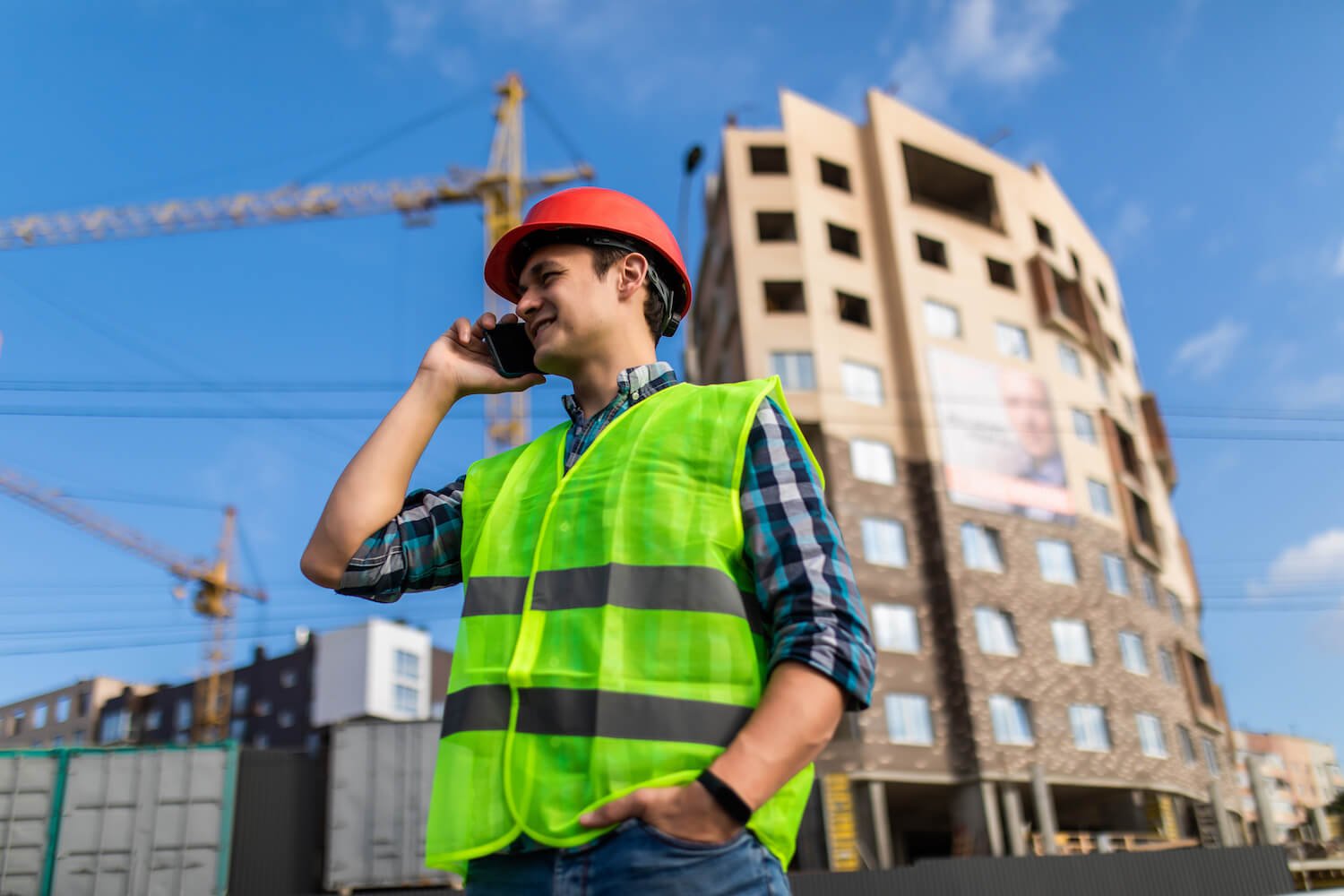 Construction worker in a yellow safety vest and red helmet, talking on a cellphone at a construction site with a building and cranes in the background.