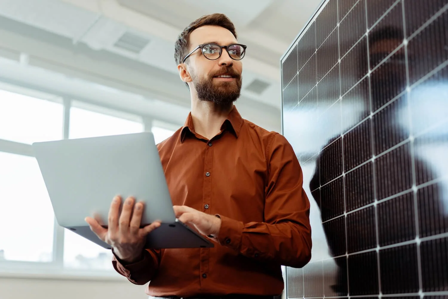A man with glasses and a beard in a brown shirt holding a laptop, standing next to a wall of solar panels inside a modern office building with large windows.