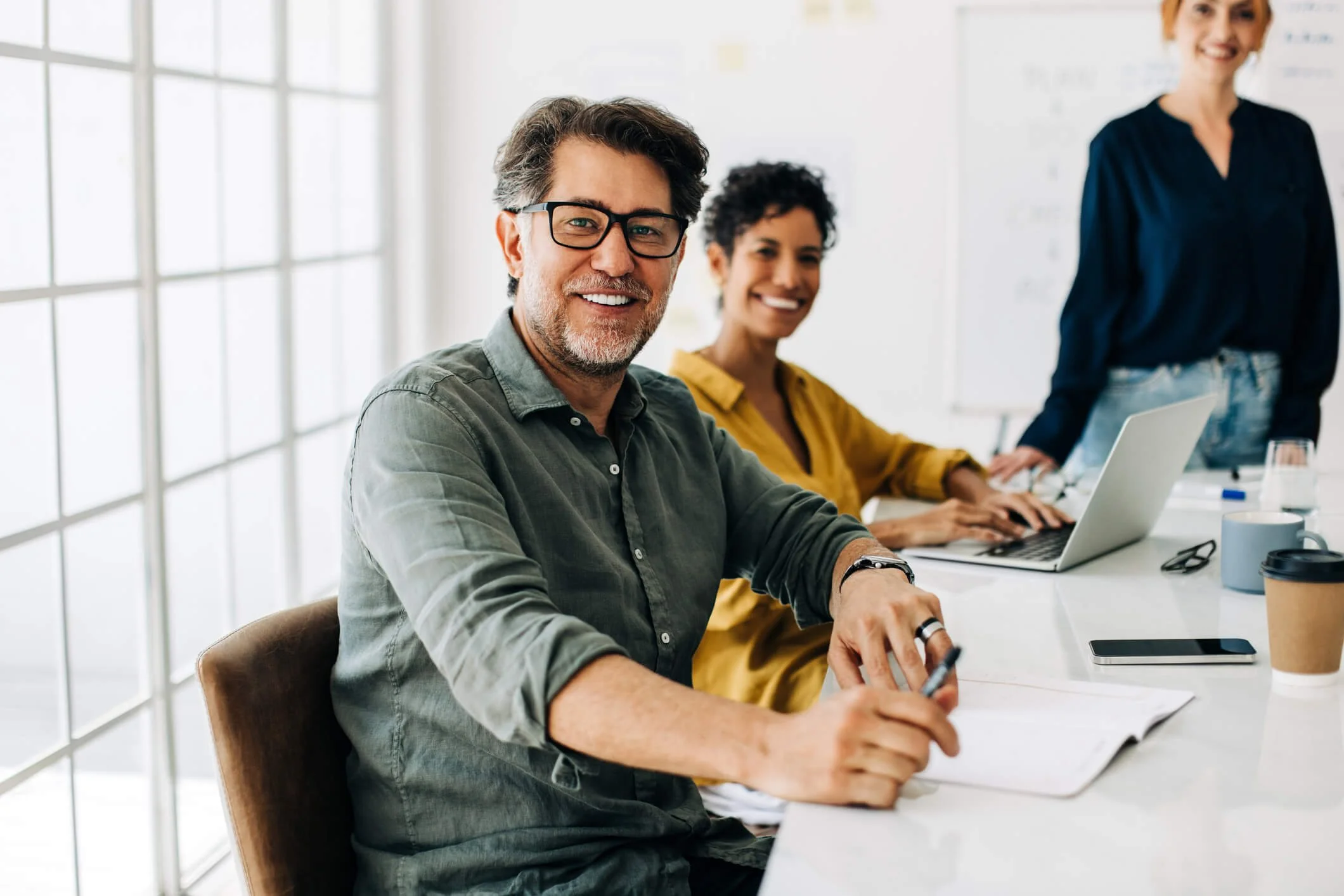 Three people sitting at a conference table in a bright office, smiling. The man in the foreground has glasses, gray hair, a beard, and is holding a pen. The woman in the middle has curly hair and is working on a laptop. The woman in the background is standing and smiling, wearing a navy blouse.