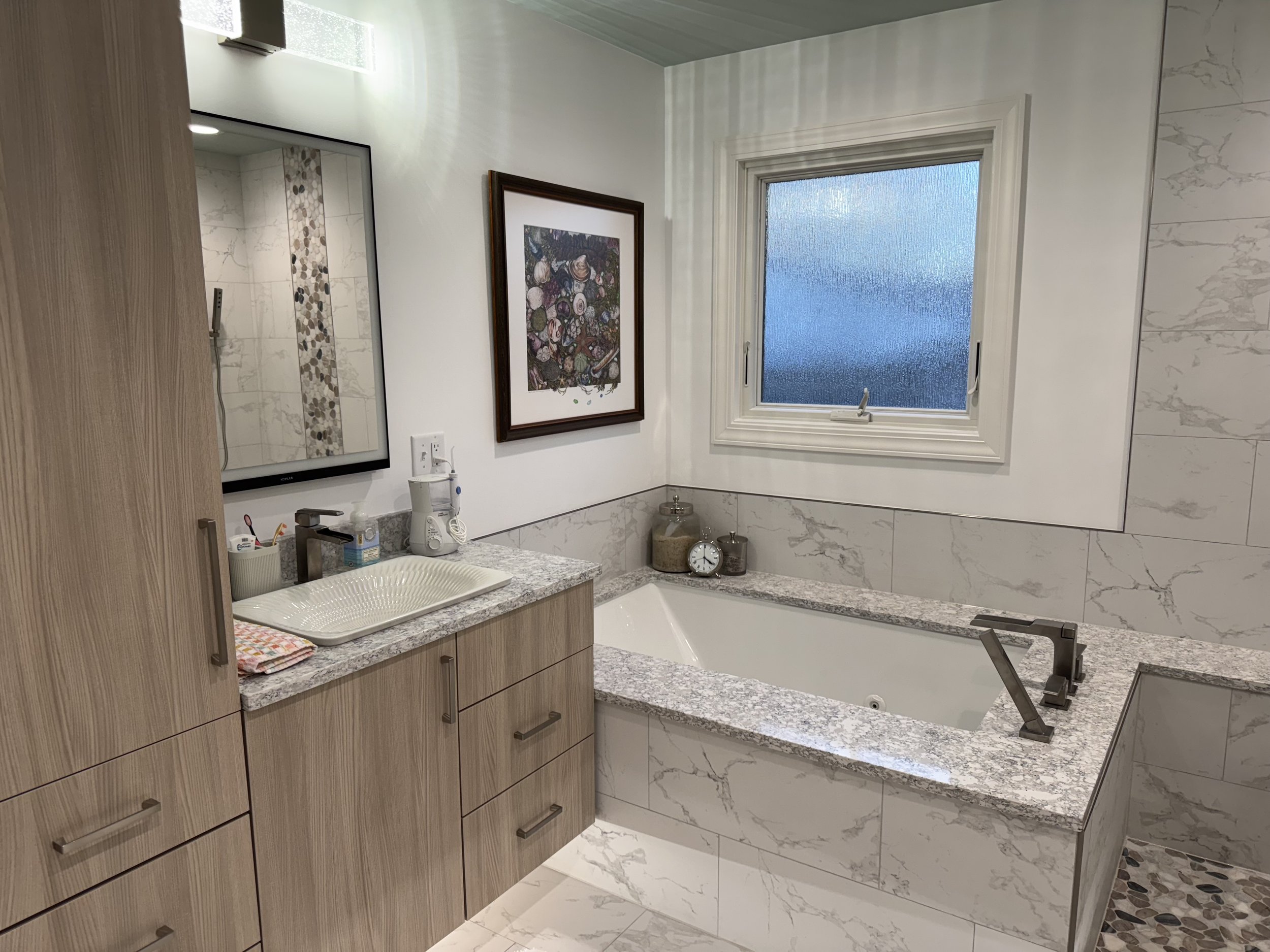 Bathroom with a bathtub, a granite countertop with a sink, a mirror, a window, framed artwork, and decorative jars.
