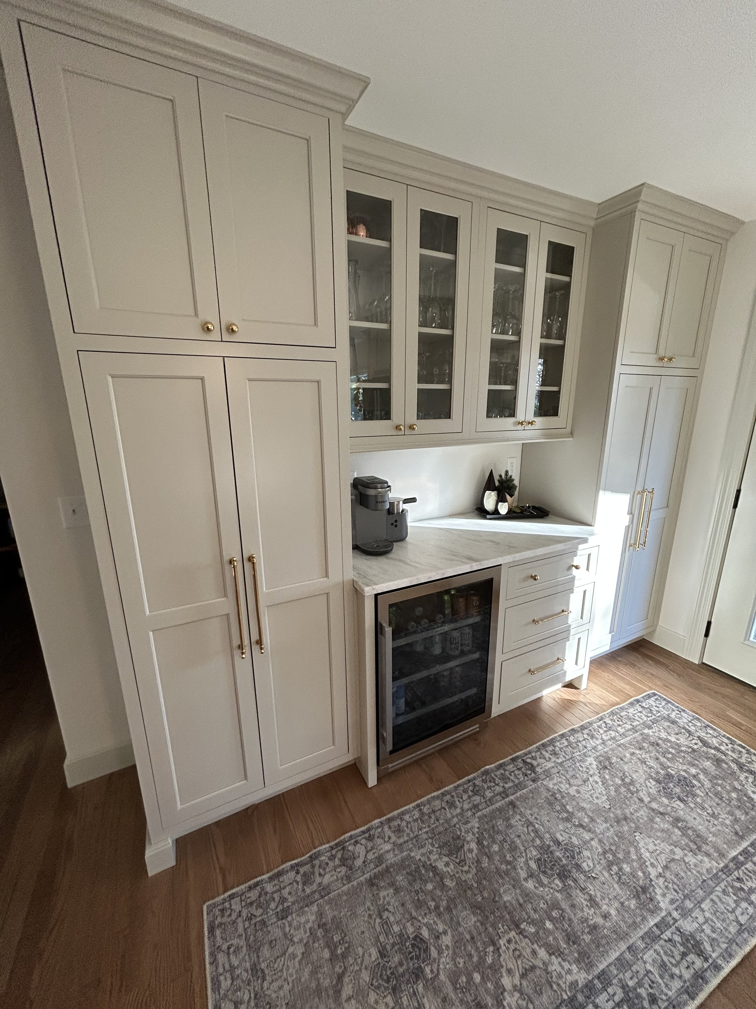 A corner of a kitchen with white cabinets, a mini wine refrigerator, and a coffee machine on the countertop. Glass-front upper cabinets display glassware. There is a decorative tray with small plants and white vases, and a patterned area rug on the hardwood floor.