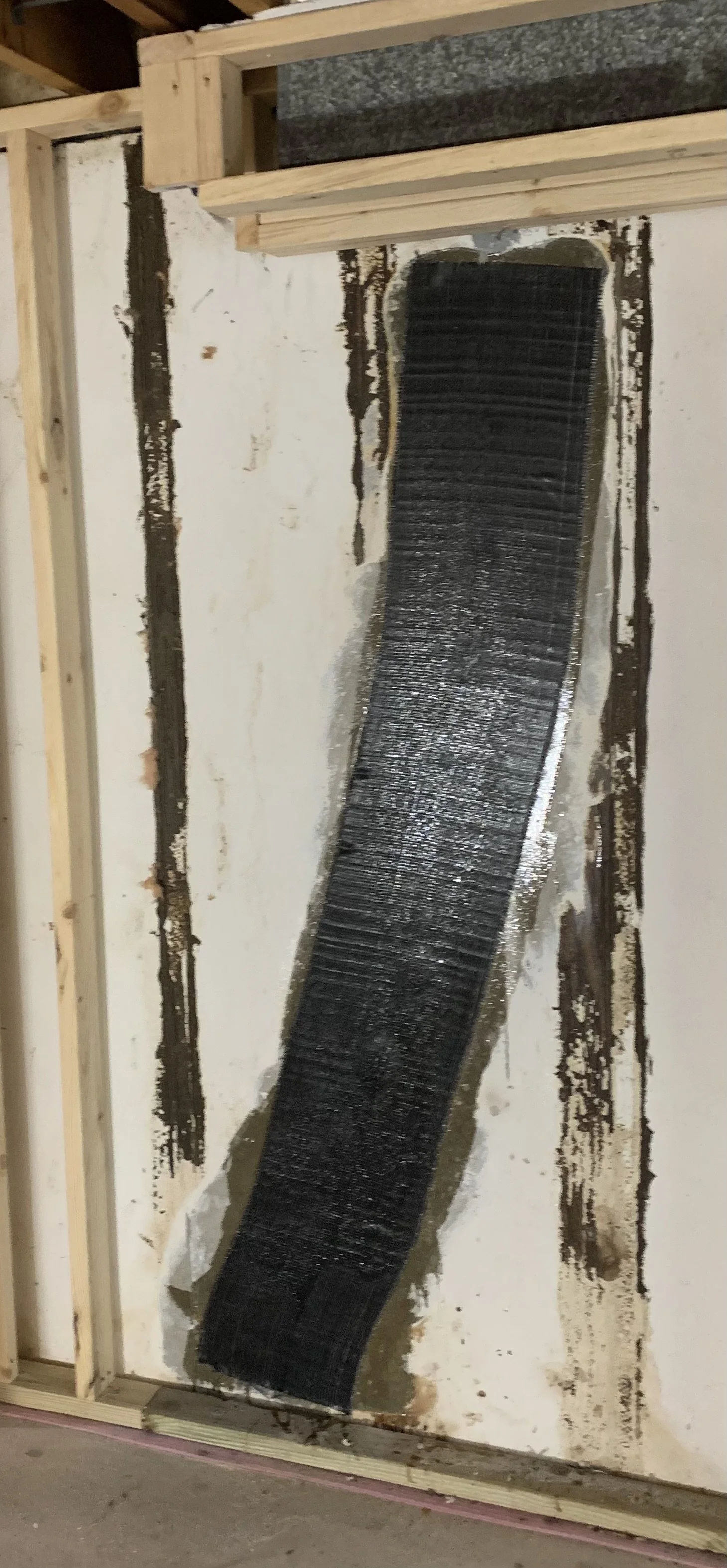 A construction site wall with black waterproofing membrane applied vertically on a section of drywall, framed with wooden studs.