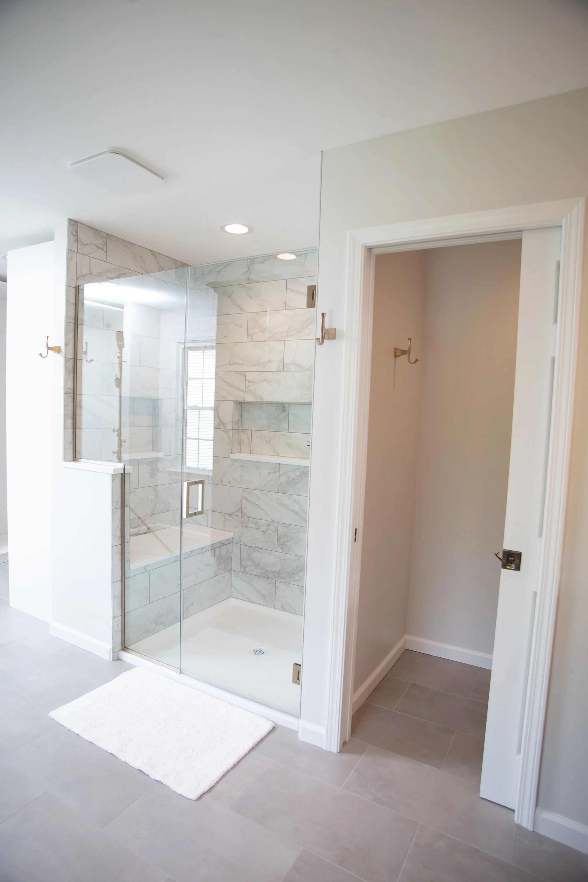 Modern bathroom with glass-enclosed marble shower, tiled walls, and a small adjacent empty closet or storage space with hooks on the wall.