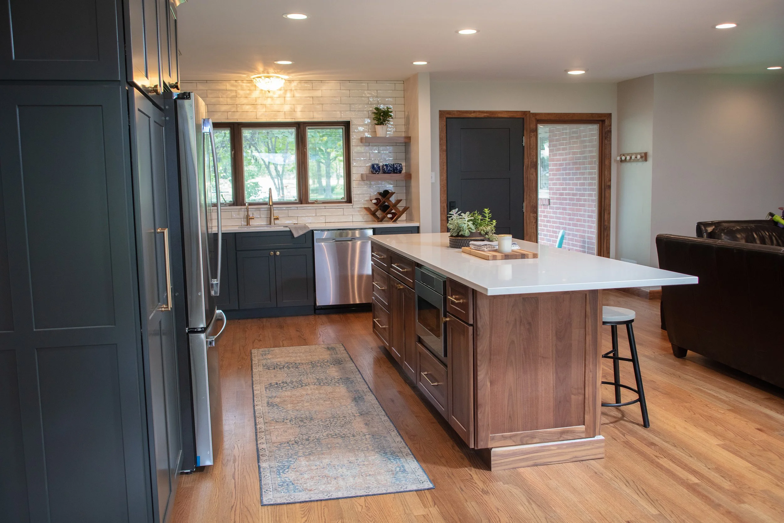 Modern kitchen with dark blue cabinets, a white island with a wooden base, and hardwood floors. There are windows above the sink, a stainless steel dishwasher, and a sliding door leading outside. The space has contemporary decor with a rug on the floor.