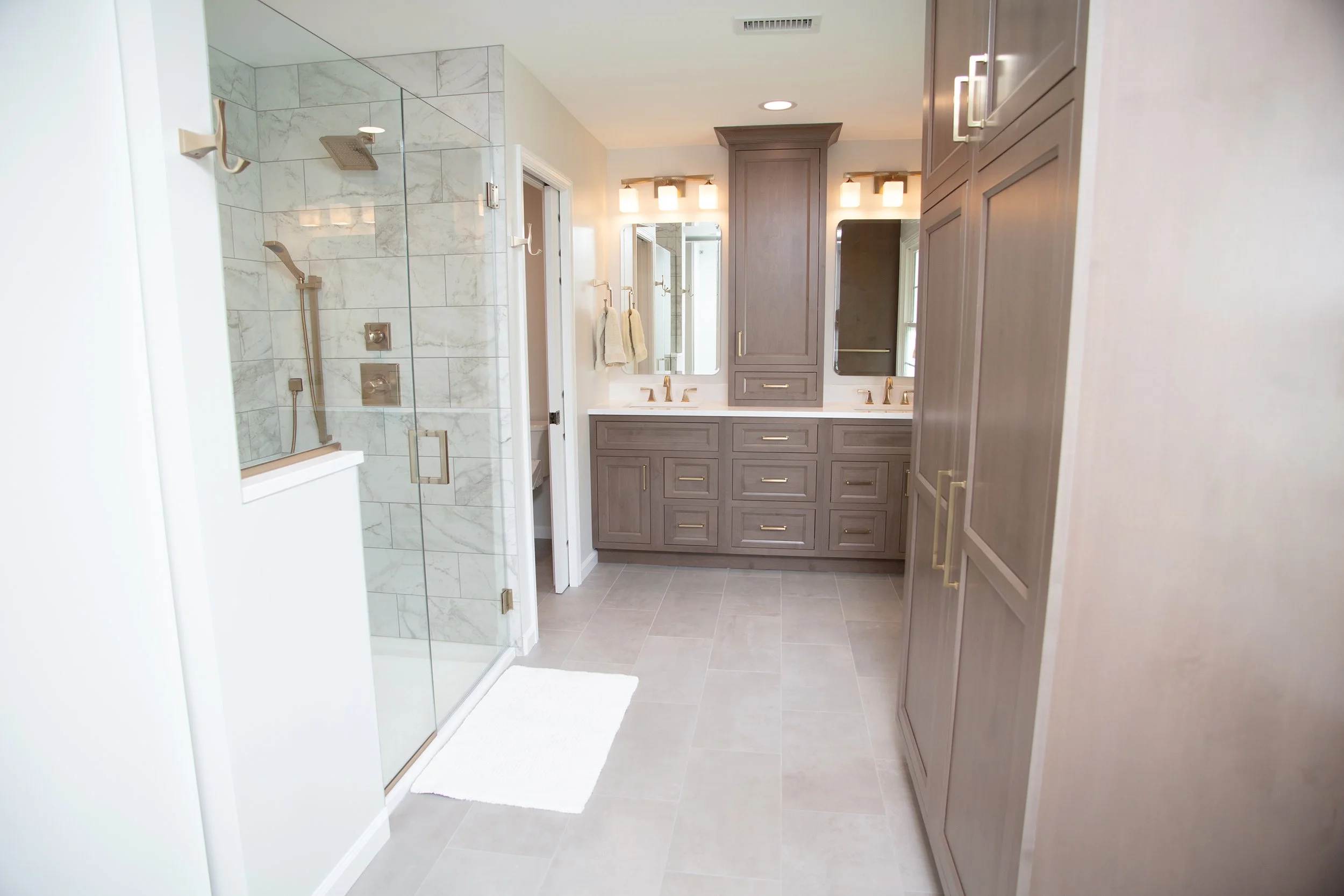 Modern bathroom with a walk-in shower with marble tiles, two sinks with mirrors and lighting, and large cabinetry for storage.