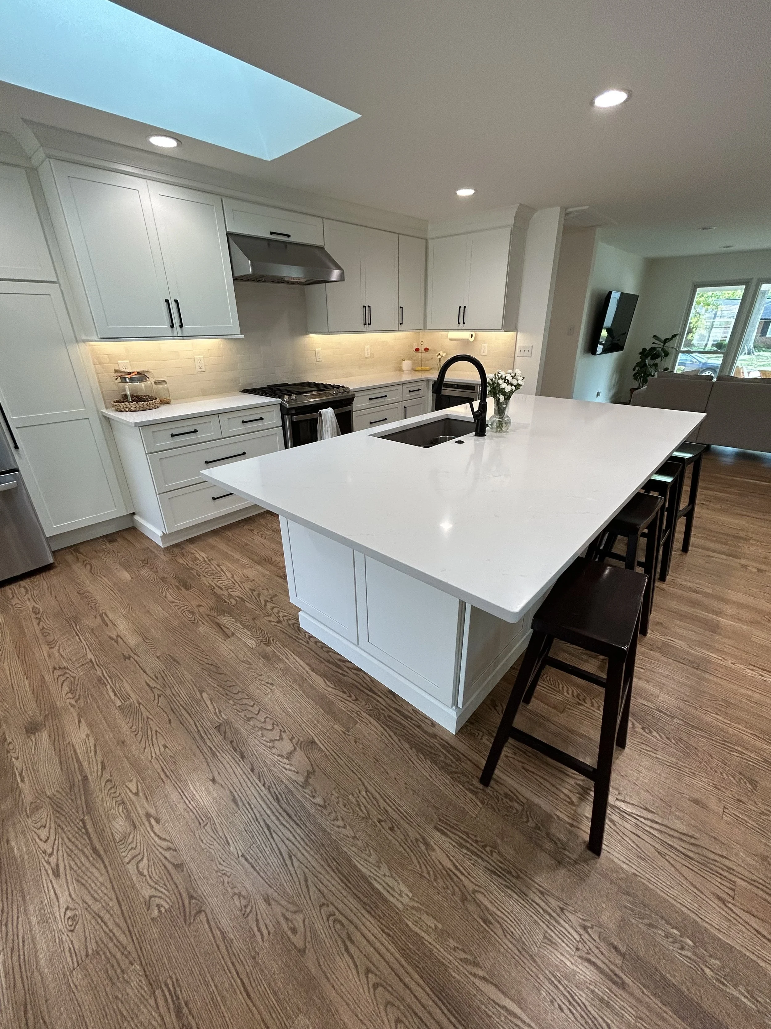 Modern kitchen with white cabinets, a large white island with a black faucet, hardwood floors, stainless steel appliances, and a ceiling skylight.