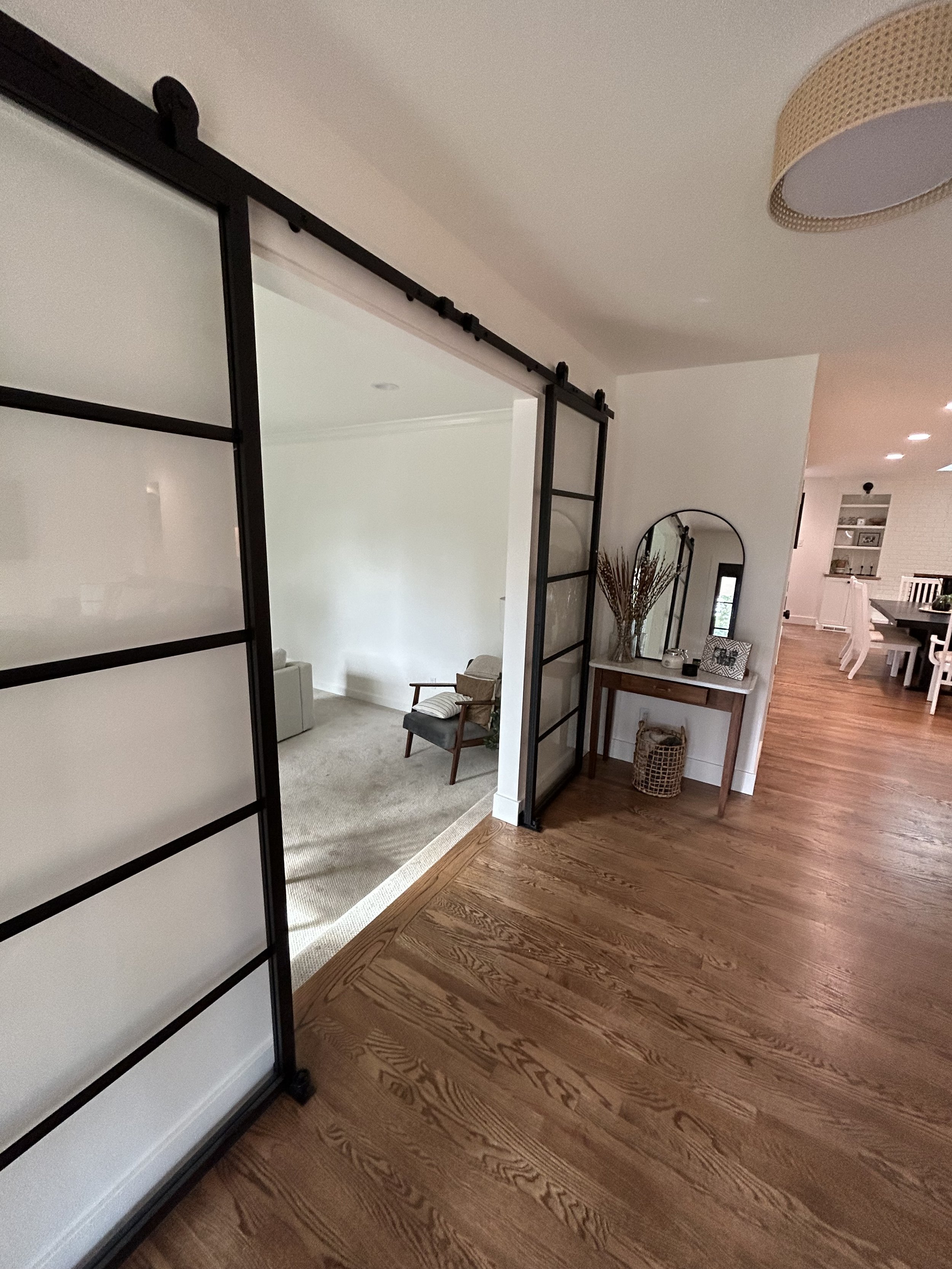 A view of a modern home interior with a wooden floor, a black-framed sliding door, a small table with decorative items, and an adjacent room with chairs and a carpeted floor.