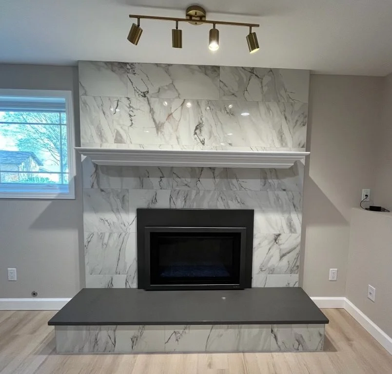 Modern living room fireplace with a black frame, set in a marble wall with gray veining, and a matching marble hearth. A white mantel shelf is above, and a brass ceiling-mounted track light with three adjustable fixtures is overhead. There is a window on the left and neutral colored walls surrounding the fireplace.