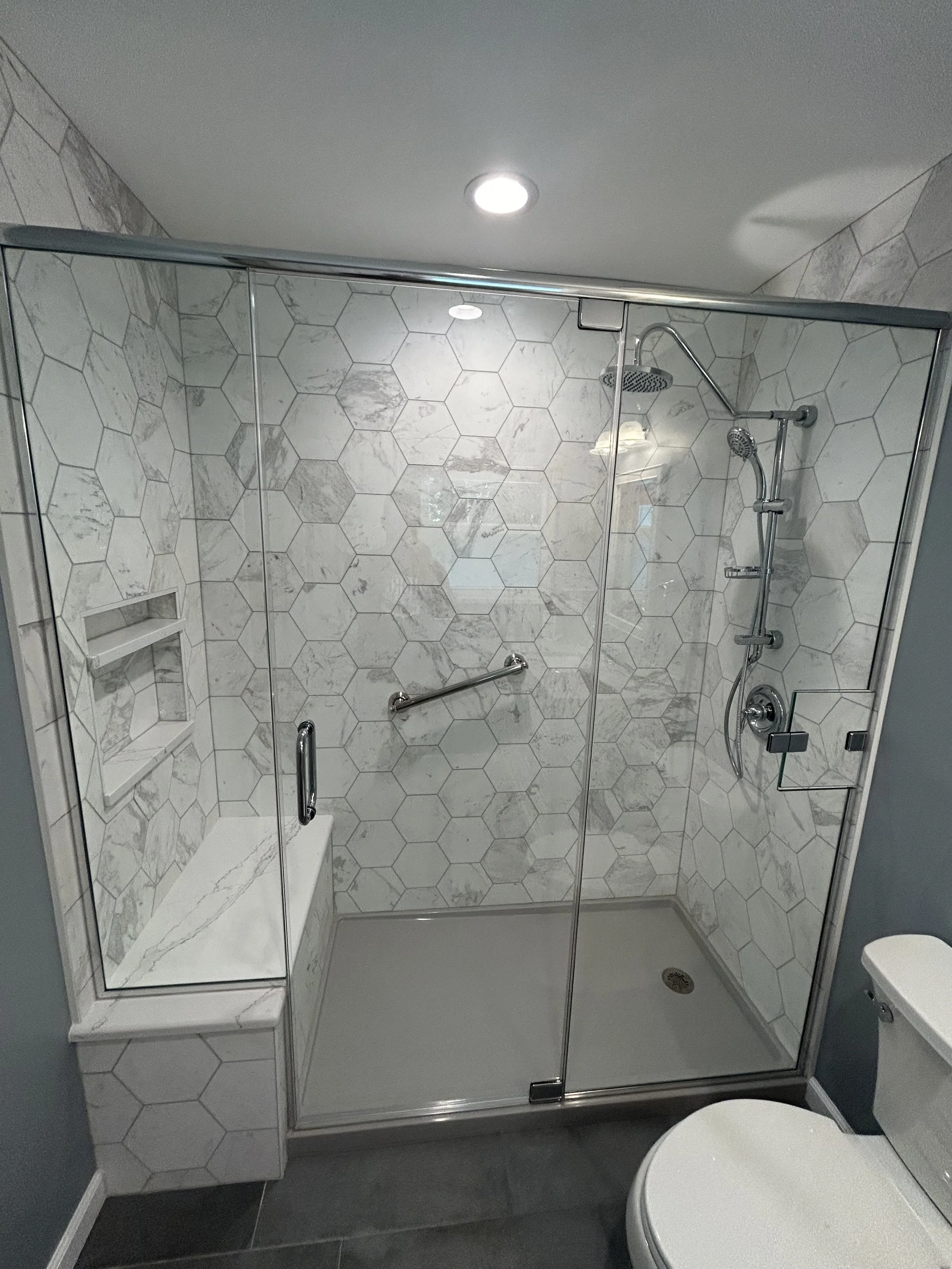 Modern bathroom with a glass-enclosed shower featuring hexagonal marble tiles, a built-in marble bench, a wall-mounted showerhead, and a handheld shower attachment, with a ceiling light and a toilet visible on the right.