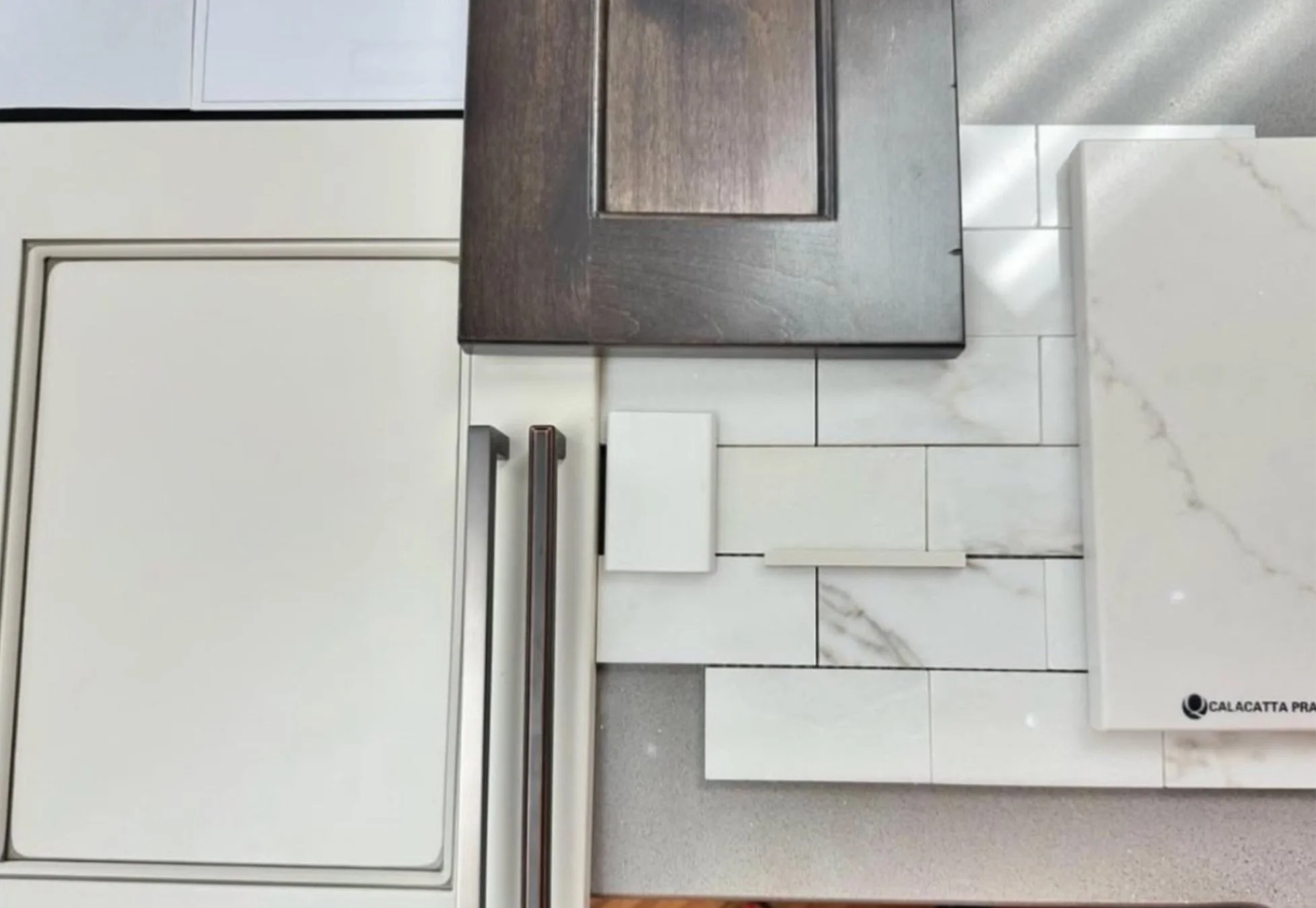 An assortment of tile samples on display, including marble, grout, and various textures, with a dark wood sample on top and a white tile with the logo 'Calacatta' on the right.