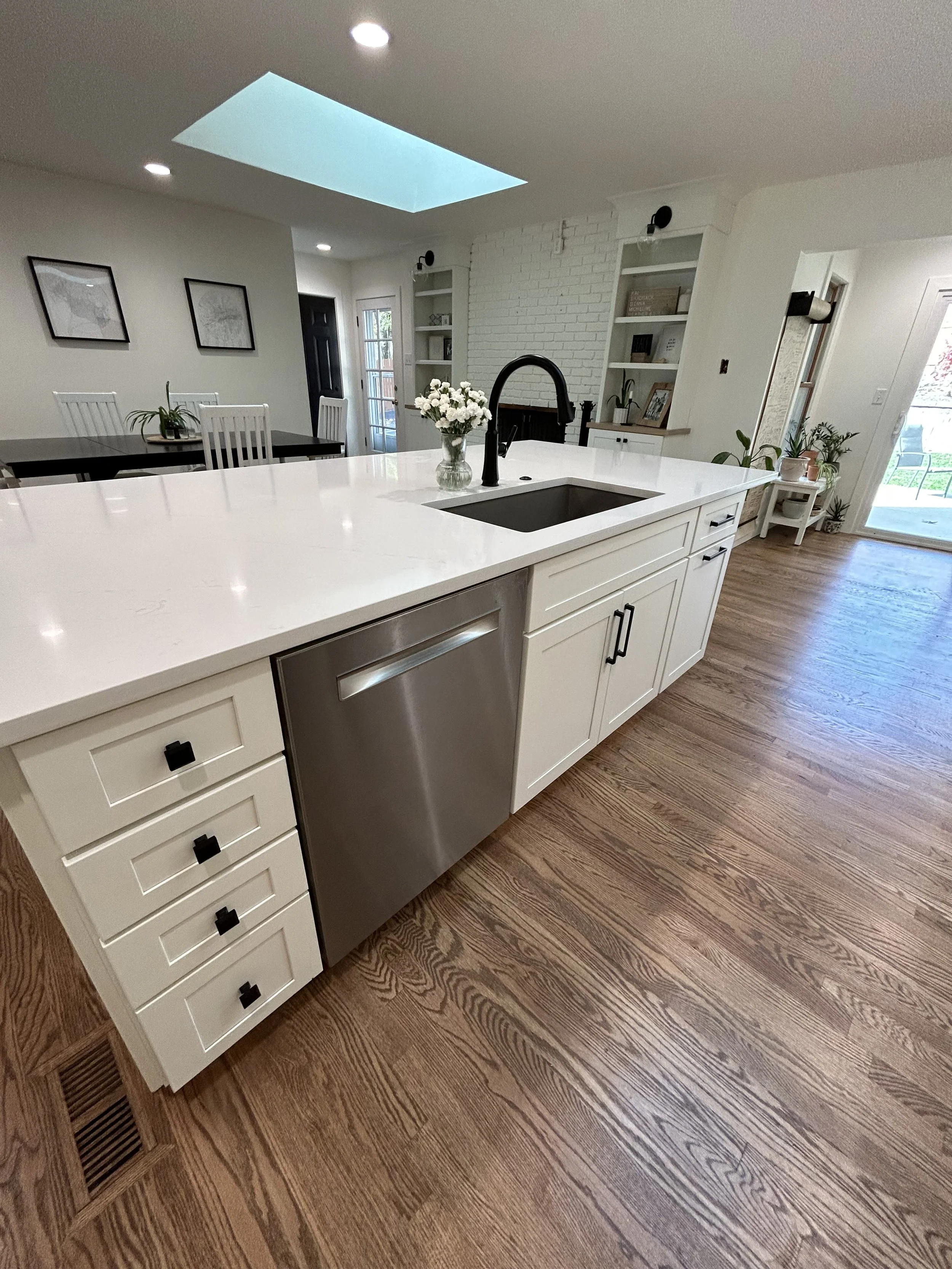 Modern kitchen with white cabinets, a large island with a white countertop, a black faucet, a vase of white flowers, wood flooring, and a skylight in the ceiling.