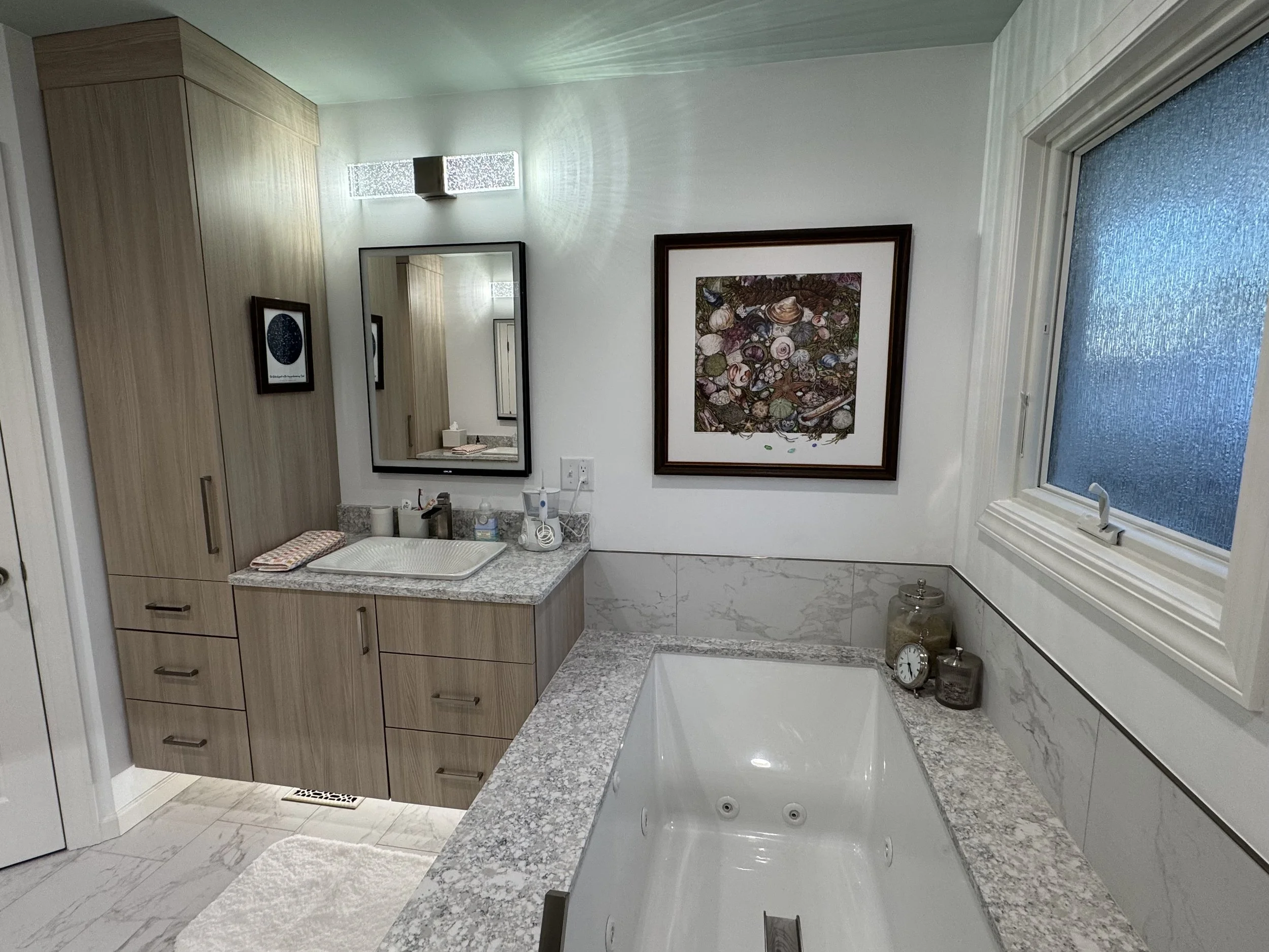 Bathroom with a large bathtub, granite countertops, wooden cabinets, a mirror, a wall art piece, a window with frosted glass, and decorative jars and a clock on the edge of the bath.