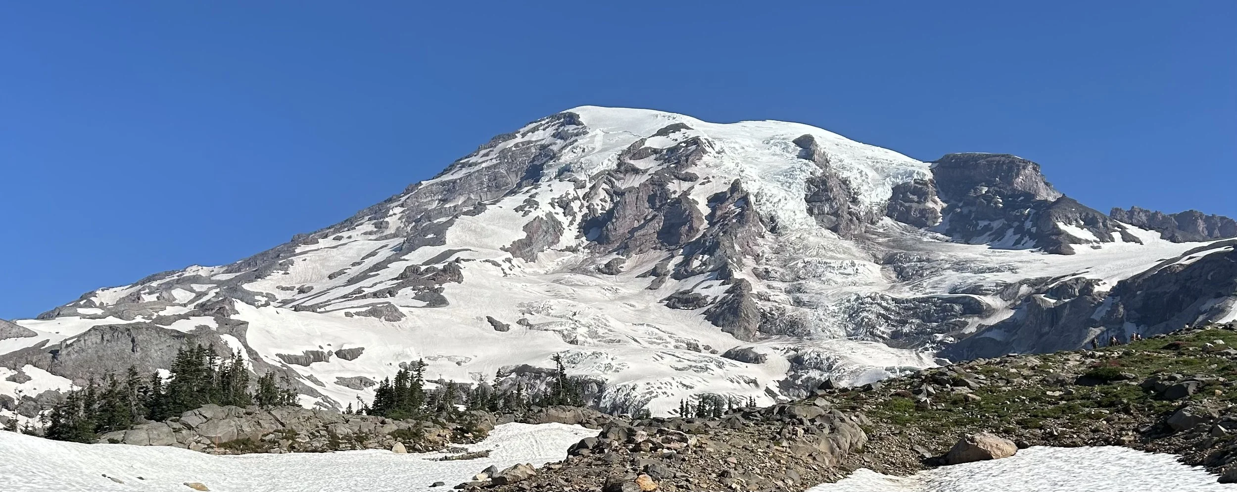 Majestic snow-capped mountain under clear blue sky with rocky and icy terrain at the base and sparse trees.
