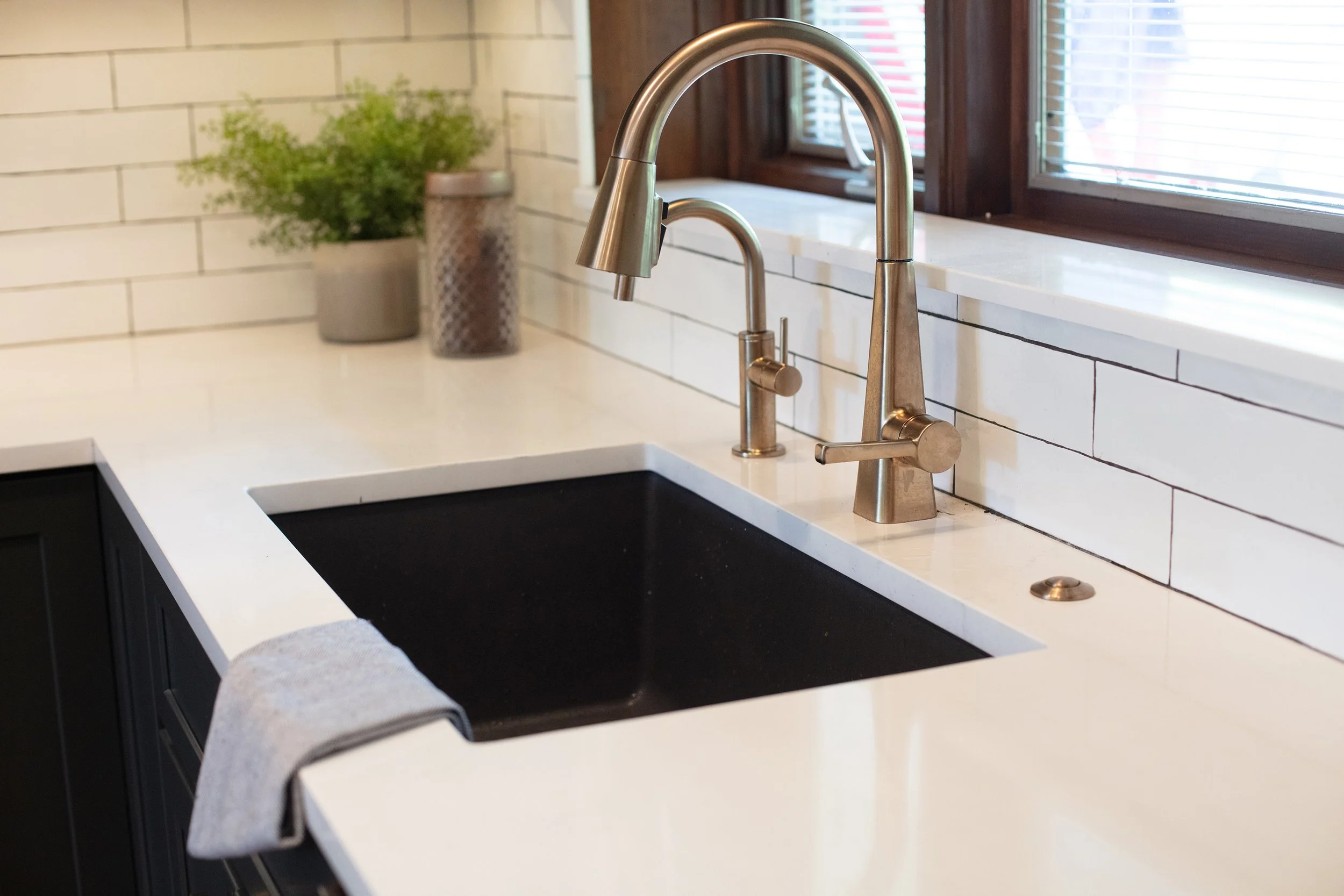 Kitchen sink with stainless steel faucet, a white countertop, and white subway tile backsplash. Two potted plants on the counter above the sink and a window with blinds.