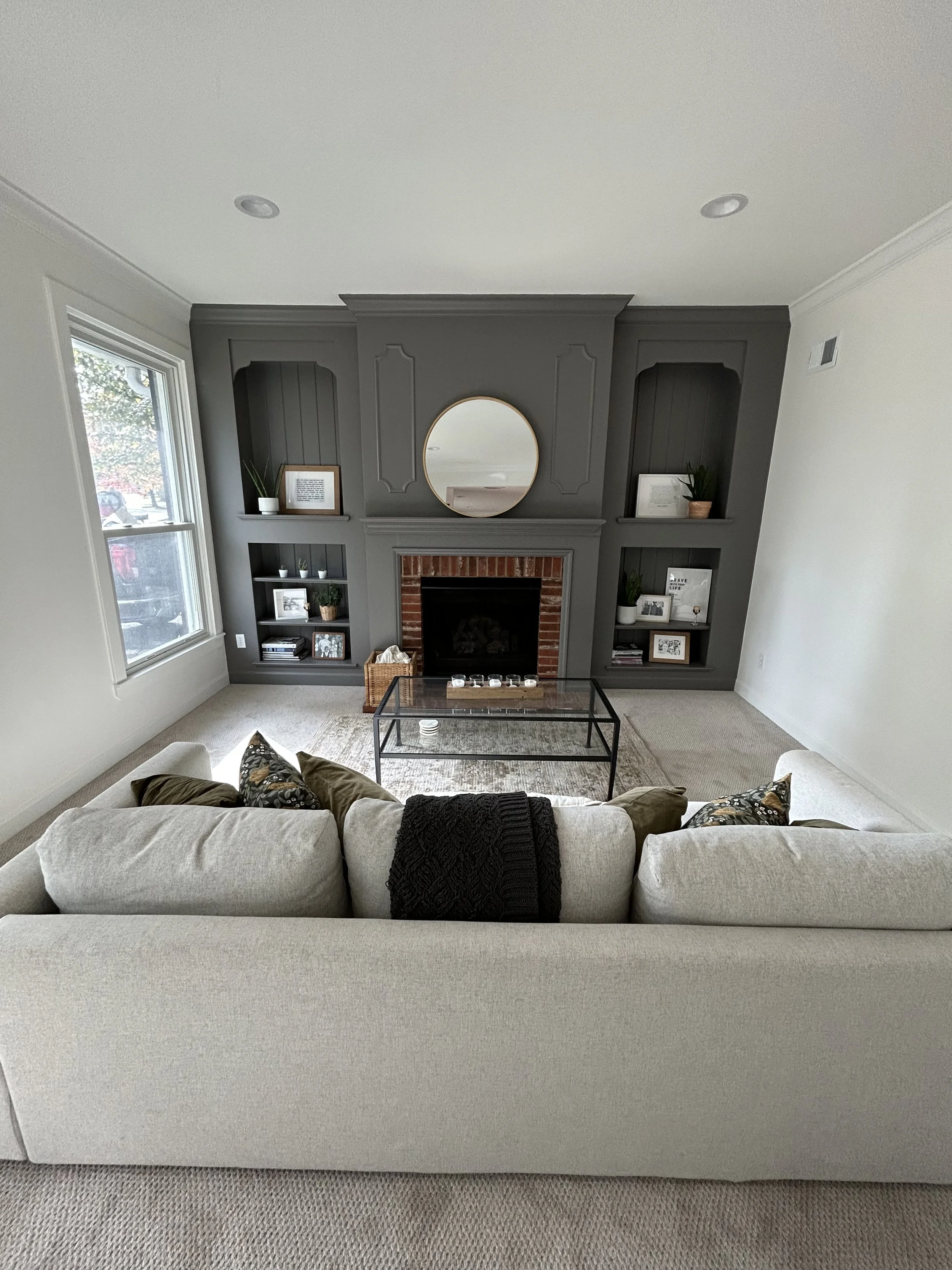 Living room with a white sofa, black metal coffee table, gray fireplace surrounded by dark gray built-in shelves, and a large window on the left, with beige carpeted floor and light-colored walls.