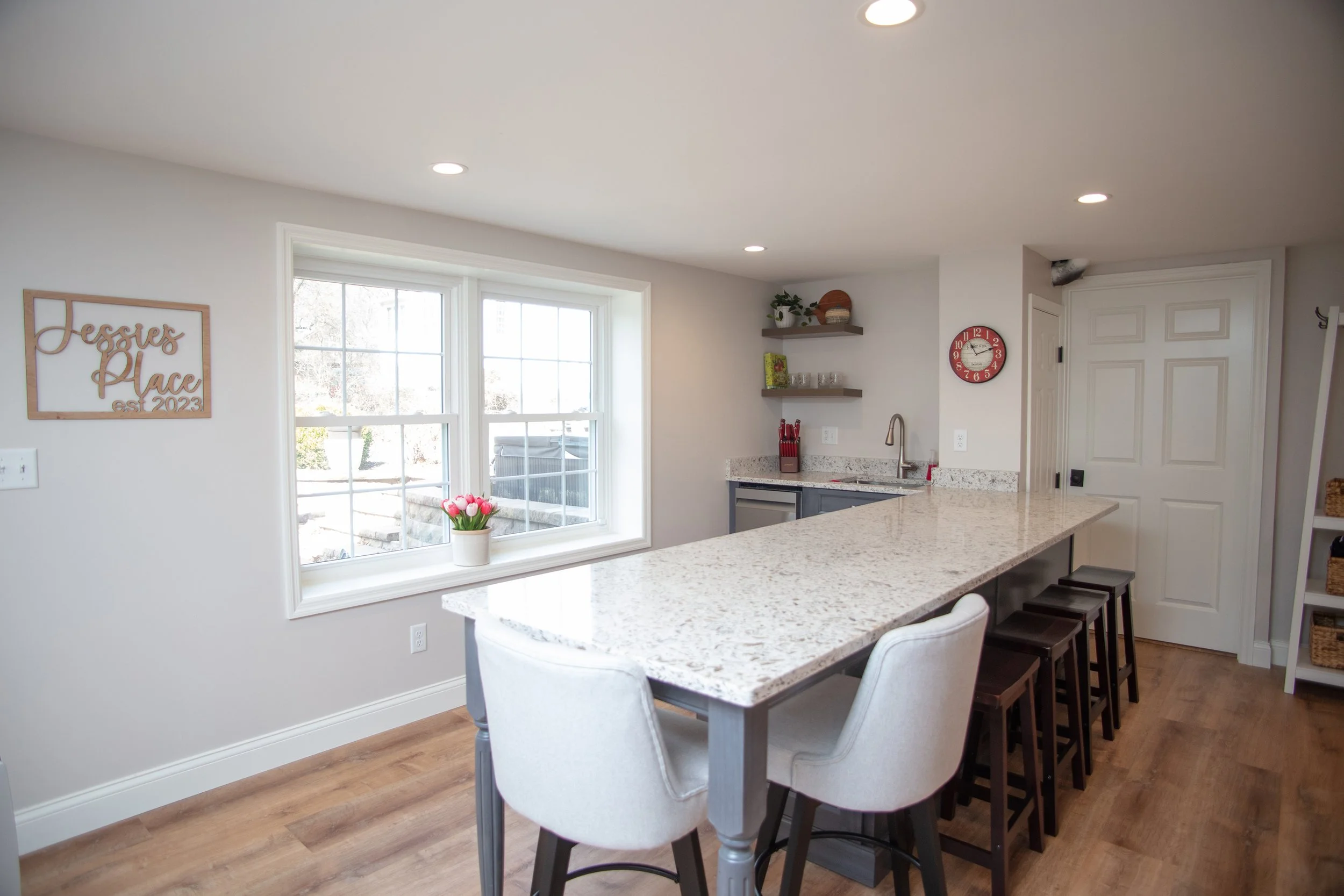 Modern kitchen with a granite island, white chairs, and wooden bar stools, featuring two large windows, a small sink area, open shelving with decor and glassware, a red wall clock, a sign that reads "Jessie's Place est 2023," and a wooden floor.