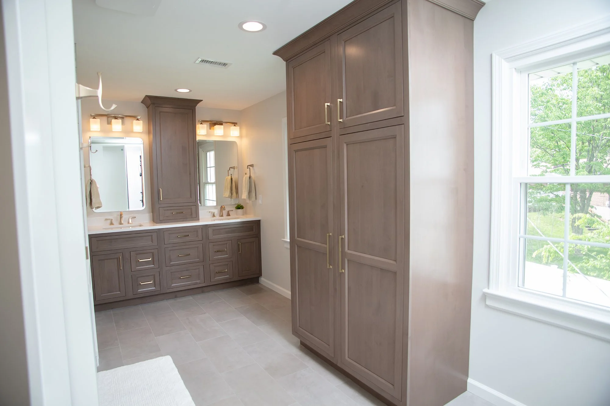 Bright bathroom with double vanity, large mirror, wall-mounted lights, wooden cabinets, and a large window showing green trees outside.