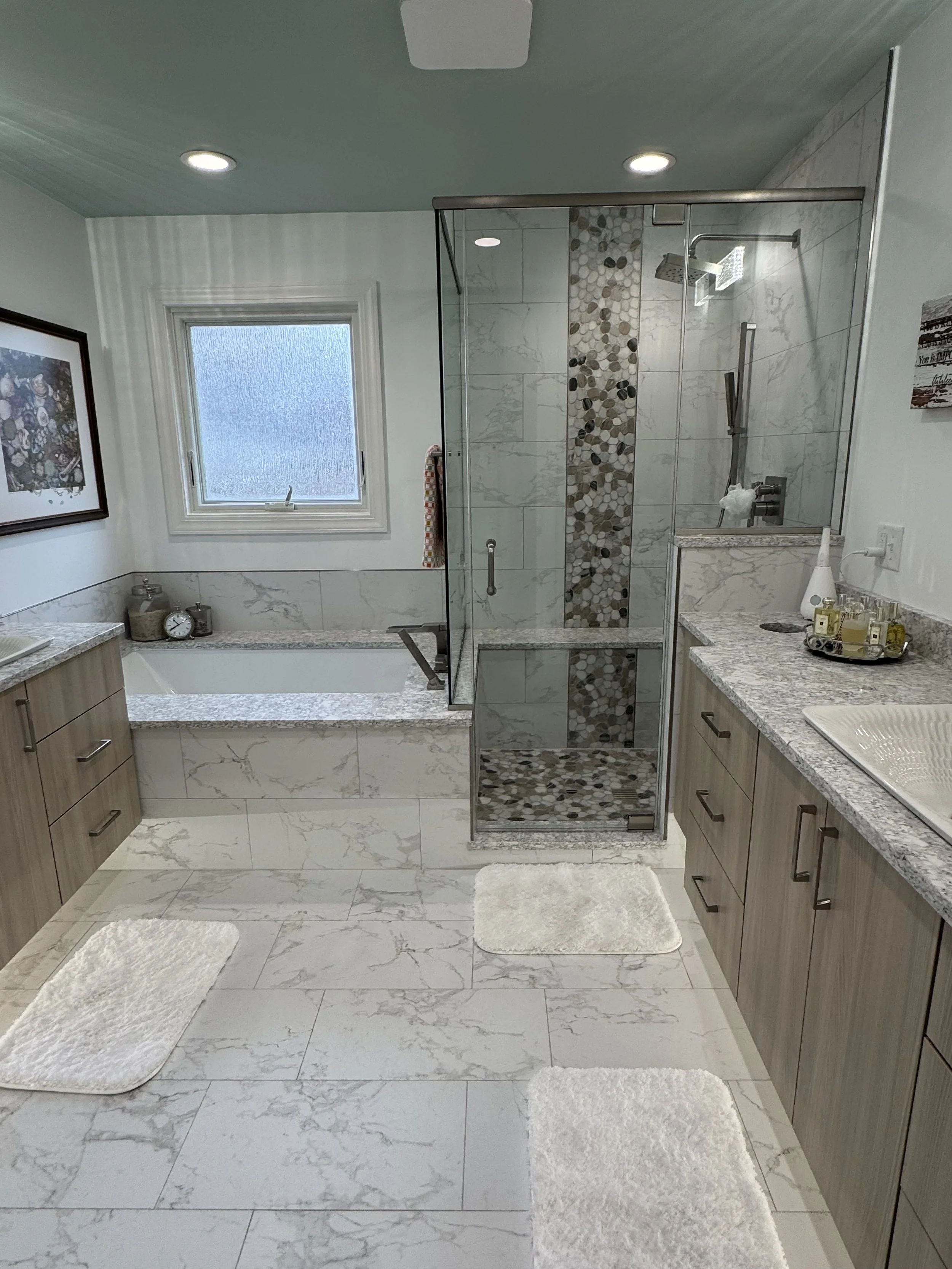 A modern bathroom featuring a bathtub, a glass shower enclosure, wooden cabinetry, a marble countertop with a sink, and white rugs on a marble floor. There is a window with frosted glass, and framed artwork on the wall.