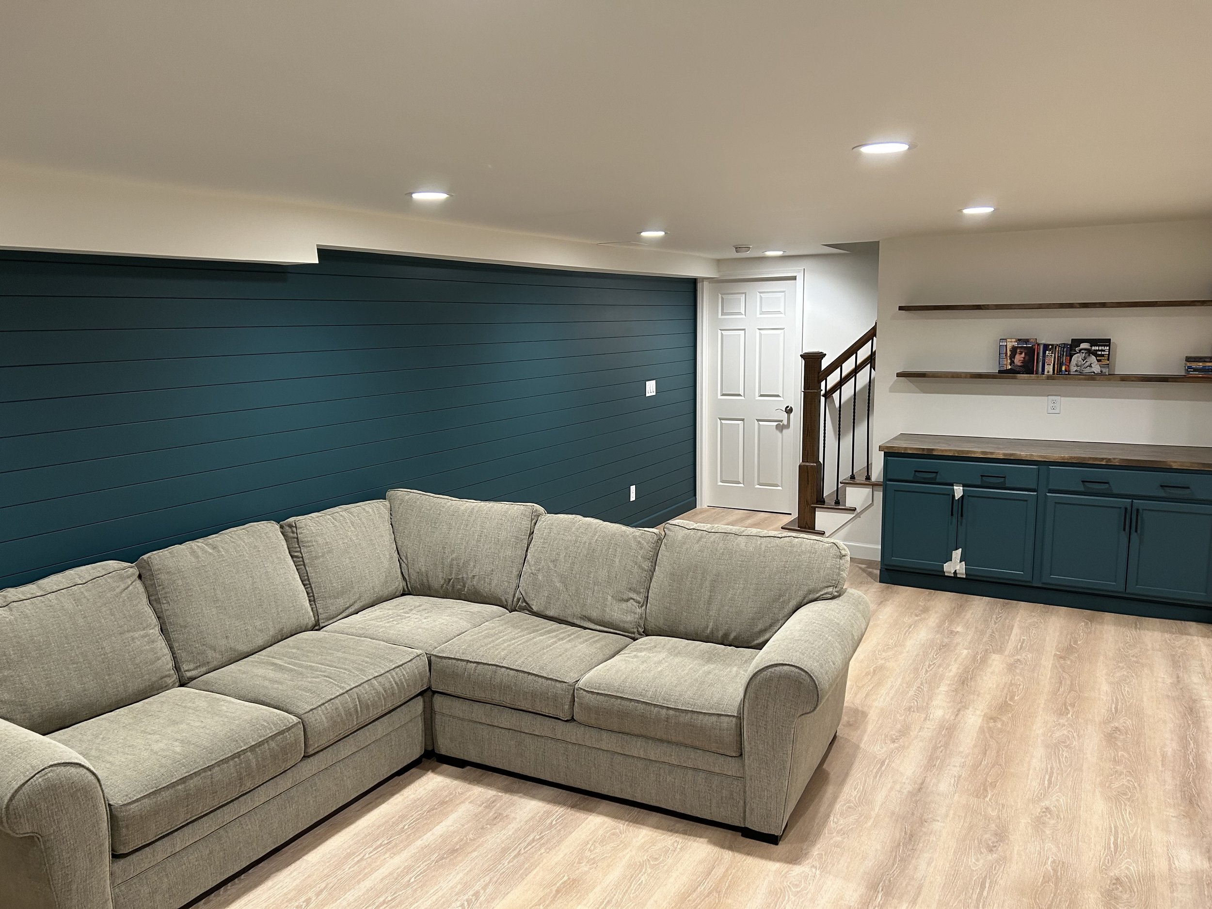 Living room with beige sectional sofa, teal accent wall, white ceiling with recessed lighting, white closet door, staircase with wooden banister, teal sideboard with wooden countertop, and two floating shelves with books and decorative items.