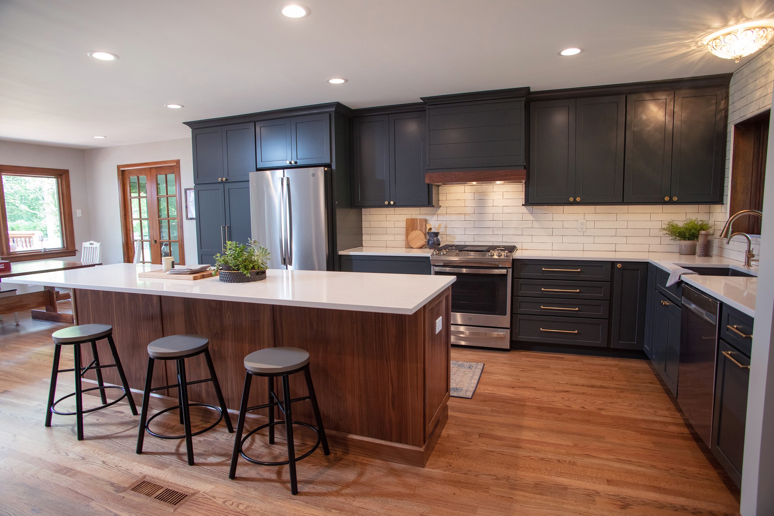 Modern kitchen with black cabinets, a white countertop island, stainless steel appliances, a white brick backsplash, hardwood flooring, and some green plants.