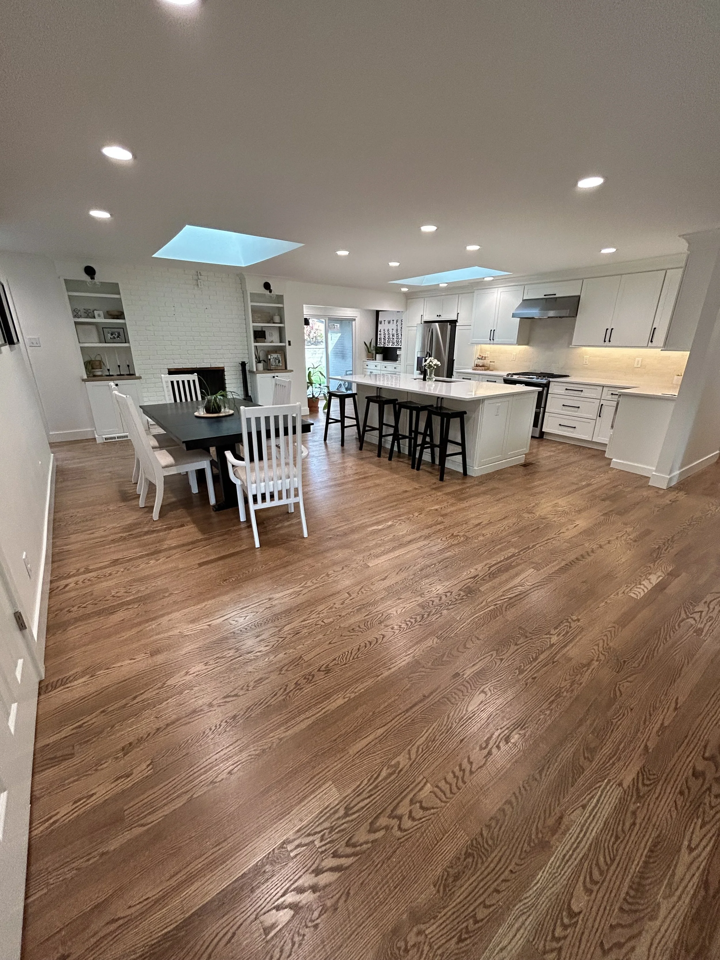 Open-concept kitchen and dining area with hardwood floors, white cabinetry, and a central island with black stools. There's a large skylight, recessed lighting, and a sliding glass door leading outside. The space includes a white brick fireplace with built-in shelves.